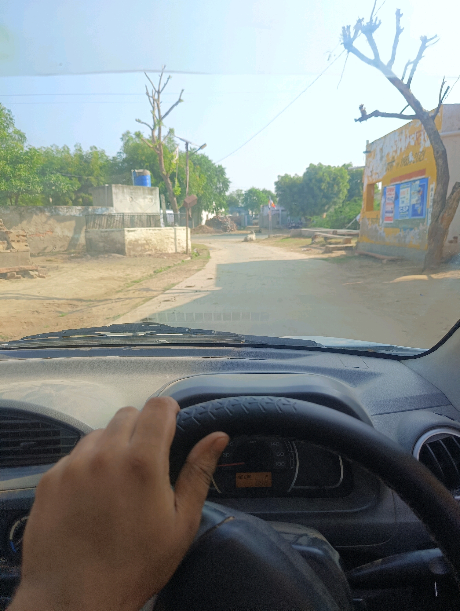 A first-person perspective shot from inside a car shows a hand on the steering wheel as the vehicle drives down a dusty road in Chhapri Bari, India. The road is lined with simple buildings and trees. The sky is clear and bright, indicating it is daytime. A dashboard with a speedometer and an orange digital display is visible in the foreground.