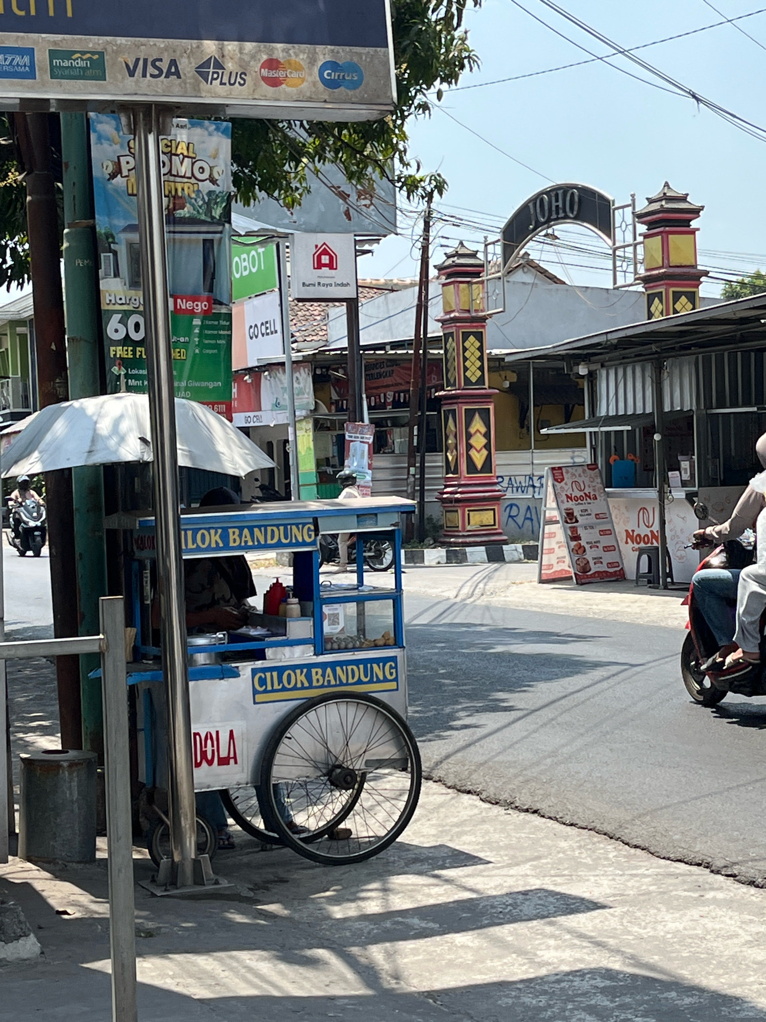This is a photograph of a street scene in Banguntapan, Indonesia, with a focus on a mobile food cart. The cart, which is blue and white, is branded with "CILOK BANDUNG" in yellow lettering. It is equipped with a white umbrella, a metal stand, and two large wheels. Inside the cart, a person is visible preparing food. Next to the cart, on the sidewalk, is a metal trash can. In the background, a road is visible with a motorcycle and its rider. Beyond the road, there are buildings, including a structure with an arched gateway that reads "JOHO". Signs for various businesses, including "GO CELL" and "NooNa", are also present. The overall environment suggests a commercial area with outdoor vendors.