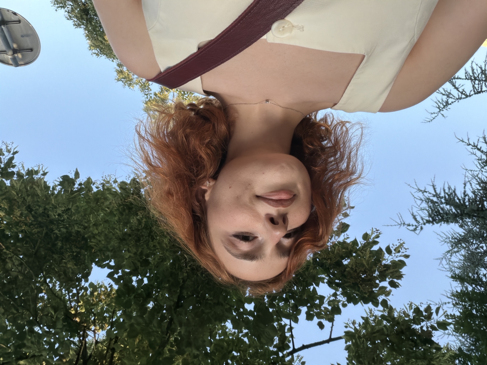 A young woman with red hair, wearing a light-colored top with a maroon strap and a small diamond necklace, looks up at the camera, which is held from a low angle, pointing upwards. Her head is tilted and her gaze is directed towards the lens, with a slight smile on her lips. Her hair is wavy and frames her face. The background consists of a clear blue sky interspersed with green foliage from trees. The image appears to be taken outdoors during the daytime, likely on a sunny day, given the bright sky. The perspective suggests the photo was taken while looking up towards the sky, possibly in a park or natural setting. There are no visible signs of text or any specific event taking place, other than a self-portrait. The overall mood is casual and perhaps a bit playful due to the unusual angle.