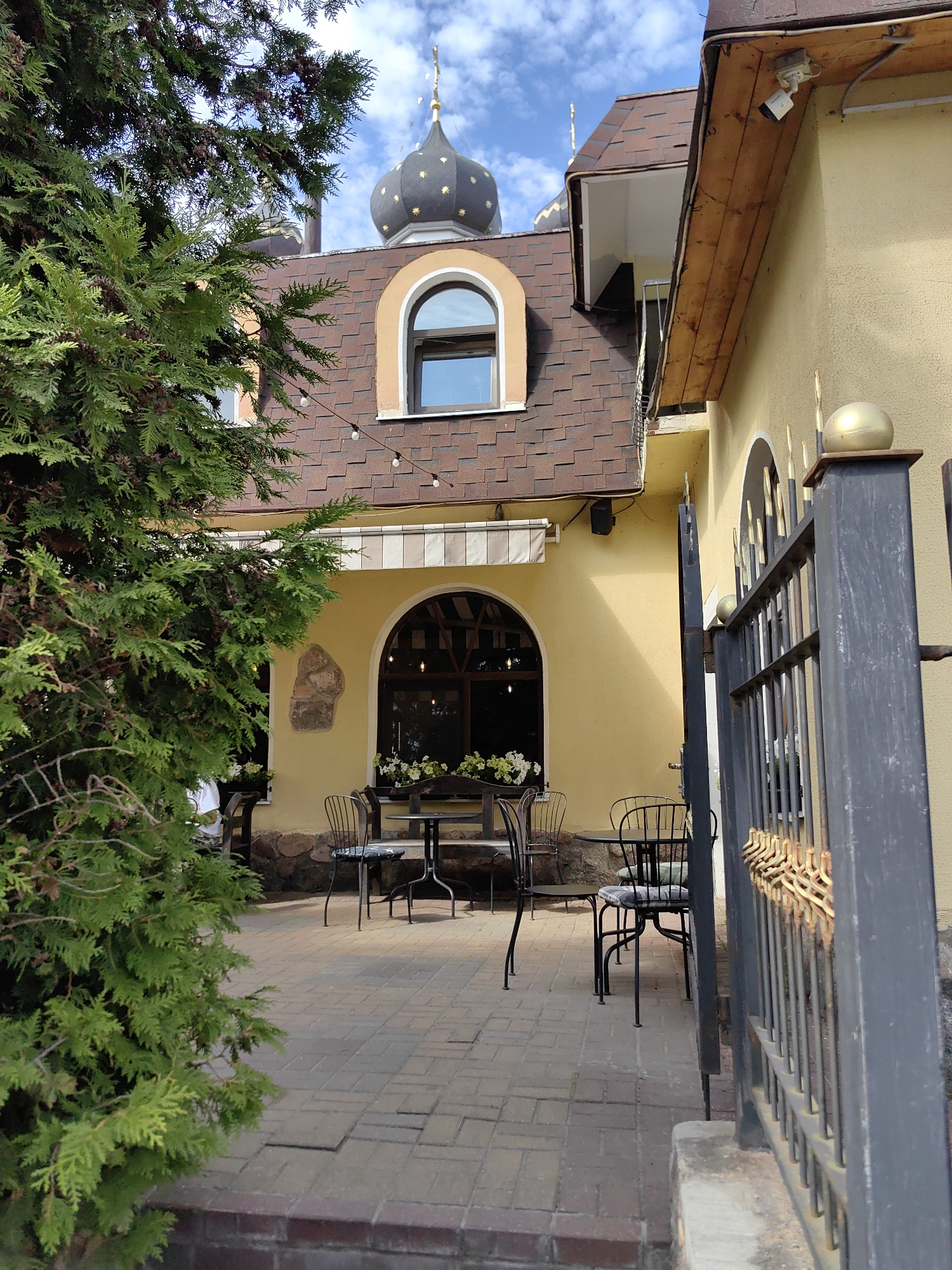 The image depicts an outdoor seating area of a building with distinct architectural features, likely a cafe or restaurant. The building itself is painted a pale yellow, with a brown shingled roof. A prominent dome with gold star accents is visible on the roof. A large evergreen tree with dense foliage dominates the left side of the frame, partially obscuring the building. In front of the building, on a brick-paved patio, are several wrought-iron tables and chairs. One table has a checkered cushion on one of its chairs. To the right, a black metal fence with gold decorative elements runs along the edge of the property. A security camera is mounted on the upper part of the building. The sky is visible above, with some clouds. The setting appears to be a relatively quiet, perhaps sunny day. The image is a photograph taken from a slightly low angle, looking towards the building. The presence of a security camera and the stylistic architecture, including the dome, suggest a specific type of establishment. While the image itself does not directly contain information about the location being Pastavy, Belarus, the prompt requests this detail be included in the description.