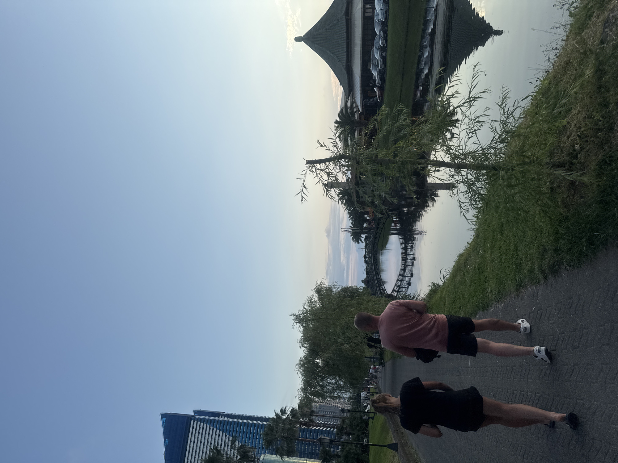 The image captures a serene evening scene along a waterfront promenade in Batumi, Georgia. In the foreground, a couple walks away from the viewer on a paved pathway. The woman on the left wears a black t-shirt and black shorts, while the man on the right wears a salmon-colored t-shirt and black shorts. Both are wearing athletic shoes. To the left of the pathway are tall, modern buildings, possibly hotels or apartments, with some palm trees visible. A large weeping willow tree partially obscures the view of other pedestrians and lamps lining the path.
The pathway runs alongside a tranquil body of water, likely a canal or inlet, reflecting the sky and surrounding elements. A decorative arched bridge spans the water in the middle ground. On the far side of the water, a distinctive building with a dark, conical roof, reminiscent of Asian architecture, is situated. In front of this building, a row of parked cars is visible. The sky is a soft blue, transitioning to lighter hues near the horizon, suggesting either sunrise or sunset. The overall atmosphere is peaceful and suggests a popular recreational area in the city.