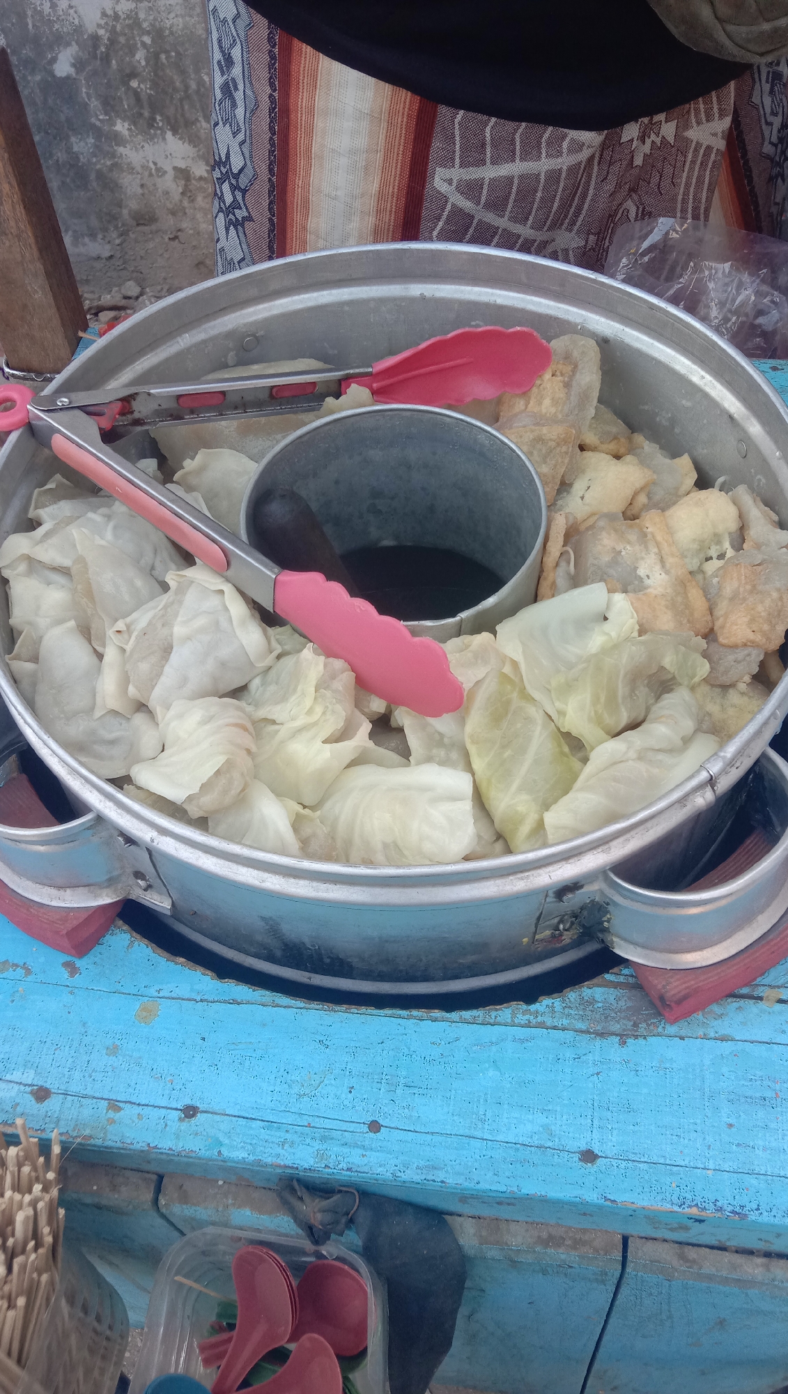 A large, circular, silver metal steaming pot sits on a weathered blue wooden surface. The pot is divided into two sections. The larger section is filled with what appears to be steamed cabbage leaves, some folded over and containing filling, and some loose leaves. A pair of pink-handled tongs rests across this section, with one tong gripping a piece of filling. The smaller section of the pot holds a dark liquid, possibly a sauce or broth, and a dark cylindrical object, possibly a pestle or ladle handle, is partially submerged in it. To the right of the steamed items, in the larger section of the pot, are pieces of fried food, appearing golden brown and somewhat irregular in shape. The blue wooden surface shows signs of wear and chipping paint, with metal rivets visible. To the bottom left of the pot, a collection of thin wooden skewers or sticks is gathered. Below the wooden surface, a clear plastic container holds several bright red plastic spoons. The background is out of focus but shows a textured grey wall on the left and a patterned fabric with stripes of brown, white, and blue on the right. The overall scene suggests a street food vendor or an outdoor food preparation area. The location is identified as Sidogiri, Indonesia.