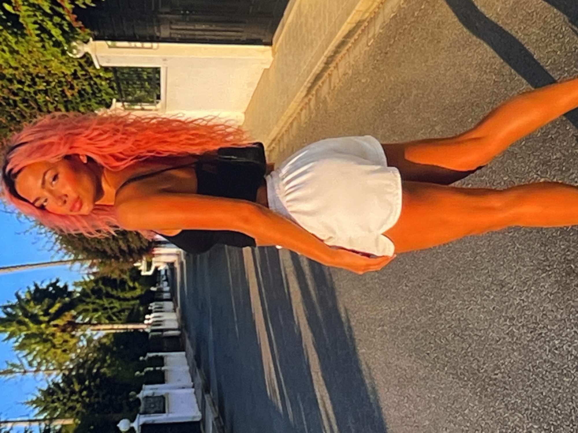 A young woman with bright pink, curly hair stands outside. She is wearing a black top and white shorts. Her skin is bronzed, and she appears to be posing for the camera. The sunlight casts long shadows on the asphalt ground, suggesting it is late afternoon or early evening. Green trees and some white structures line the background. The scene is outdoors, likely on a street or pathway. No text is visible in the image.