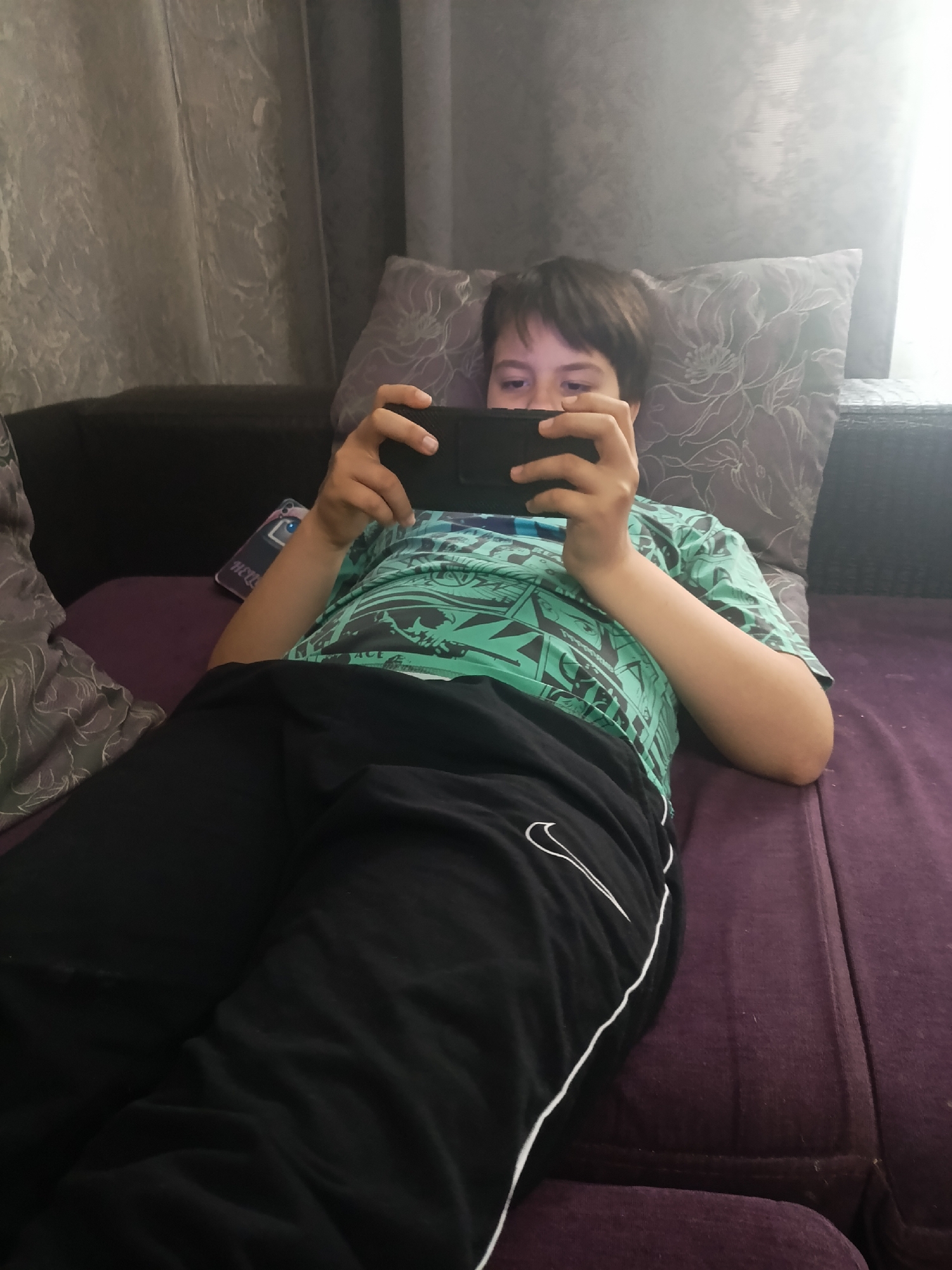 A young person lies on a purple couch, holding a smartphone and looking at it. The person is wearing a green printed t-shirt and black sweatpants with a white stripe and Nike logo. Behind the person are patterned pillows and a curtain. A phone case is visible next to the person on the couch. It is not possible to confirm the location is Krasnodar, Russia from the visual information.