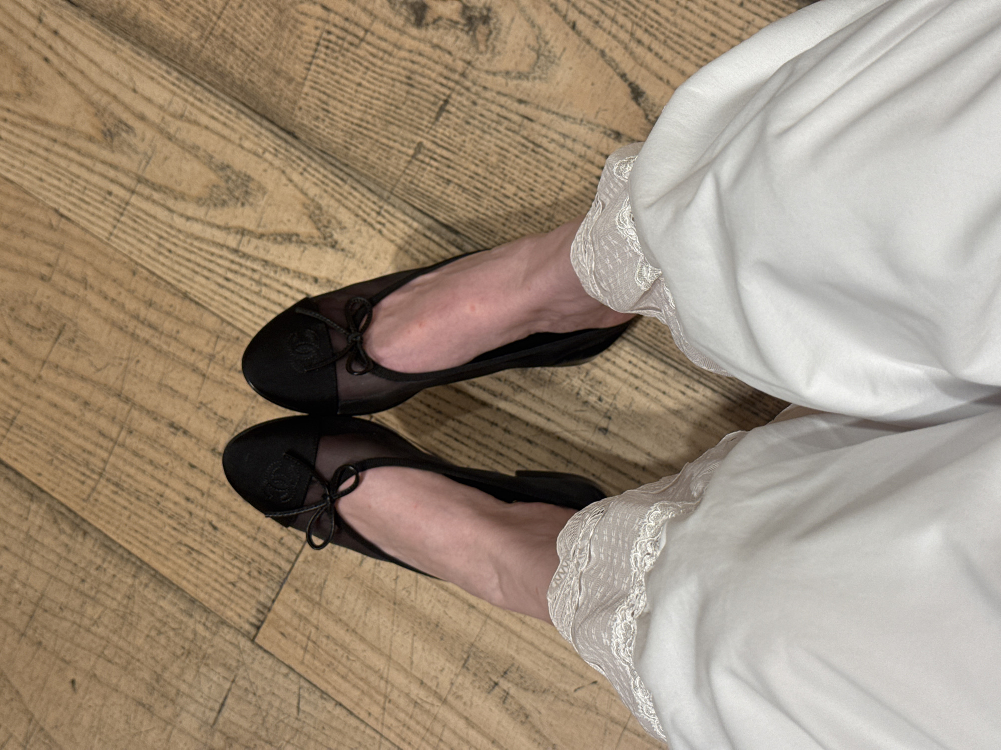 The image shows a top-down view of a person's feet wearing black ballet flats with bows on a wooden floor. The person is wearing white pants with lace trim at the bottom. The wooden floor has a light brown color with a visible wood grain pattern. The location is unknown and cannot be determined from the image.