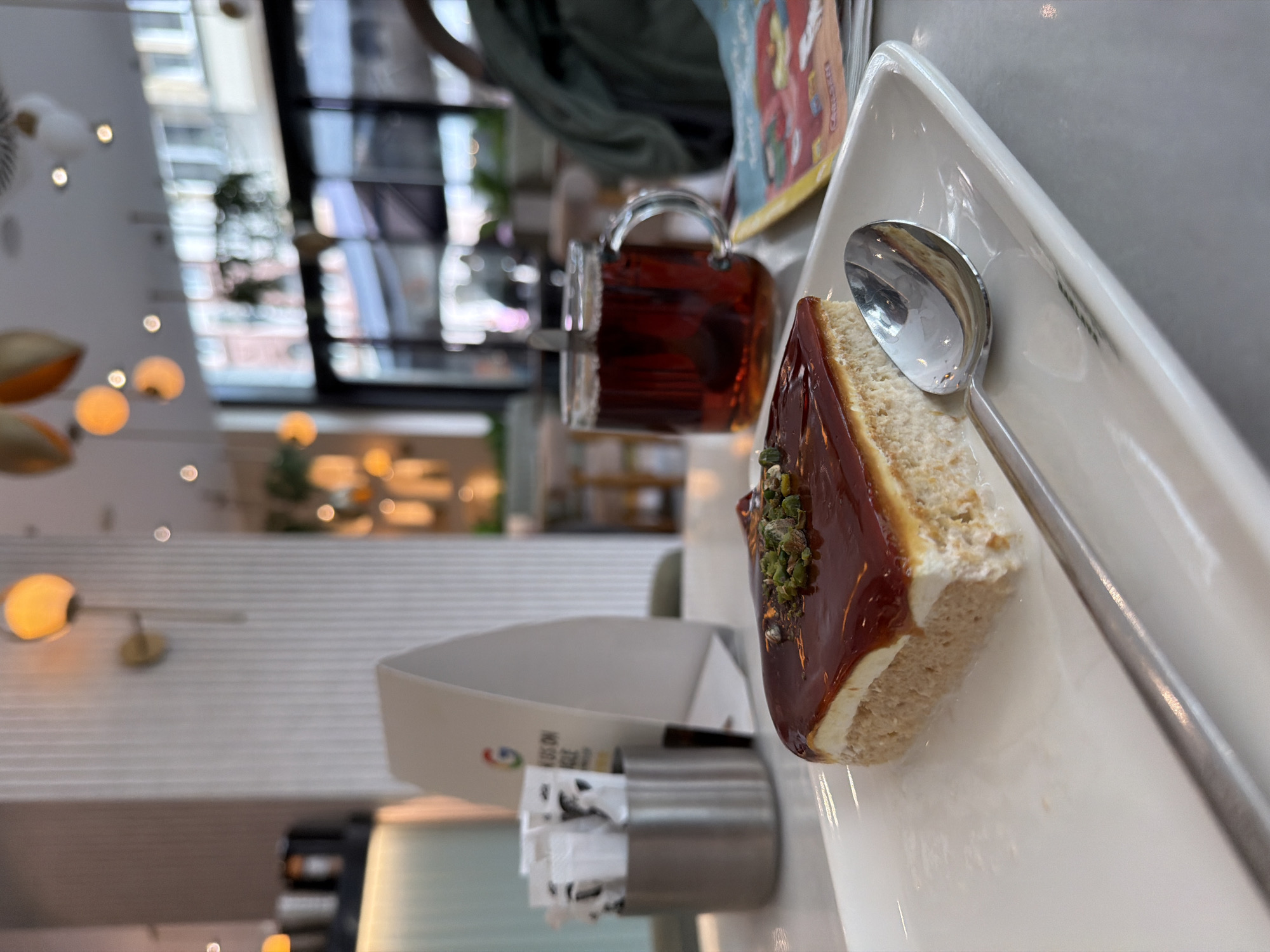 This image captures a close-up, overhead view of a dessert and beverage set in a modern cafe or restaurant, likely in Wiesbaden, Germany. The primary subject is a rectangular slice of cake, possibly a cheesecake or a similar layered dessert, placed on a white, slightly triangular plate. The cake features a light-colored base, a creamy white layer, and is topped with a glossy, dark caramel-colored glaze, adorned with green crushed pistachios. A silver spoon rests on the plate beside the cake.
Behind the plate, a clear glass mug holds a dark red liquid, resembling tea. Further back, a metal cylindrical holder contains several white sugar or condiment packets. Adjacent to this holder is a white stand or container with a colorful logo, possibly a stylized flag or emblem, and some text written in an Asian script, alongside a smaller red logo. A partially visible, brightly colored book or magazine cover lies blurred in the background, showing some English text that is difficult to discern clearly.
The setting appears contemporary, characterized by sleek white surfaces and minimalist design elements. The background is softly blurred, showing warm-toned spherical lights as decorative accents. Large windows or glass partitions are visible, reflecting blurred architectural shapes and ambient light from outside, indicating it is daytime. The scene suggests a relaxed atmosphere where someone is enjoying a sweet treat and a drink.