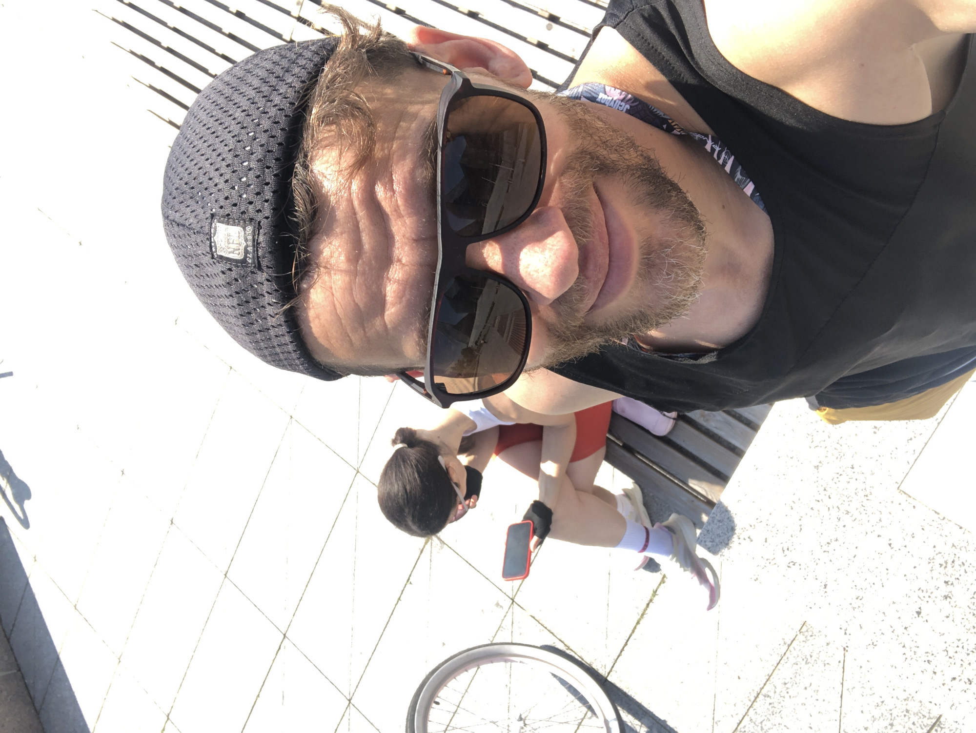 This media file shows two people outdoors on a sunny day, likely in Saint Petersburg, Russia, given the context. The main subject is a man taking a selfie, wearing a black beanie, sunglasses, and a black tank top. He has a beard and appears to be in his 30s or 40s. Behind him, a woman is sitting on a wooden bench, wearing a white top, red shorts, white socks with pink stripes, and sneakers. She is holding a red smartphone. A bicycle wheel is visible in the foreground at the bottom of the image. The ground is paved with light-colored tiles, and there are diagonal shadows cast by slats of wood, suggesting the sun is relatively low in the sky, possibly morning or late afternoon. The overall mood seems relaxed and casual, possibly indicating a break during an outdoor activity like cycling. There is no visible text in the image.