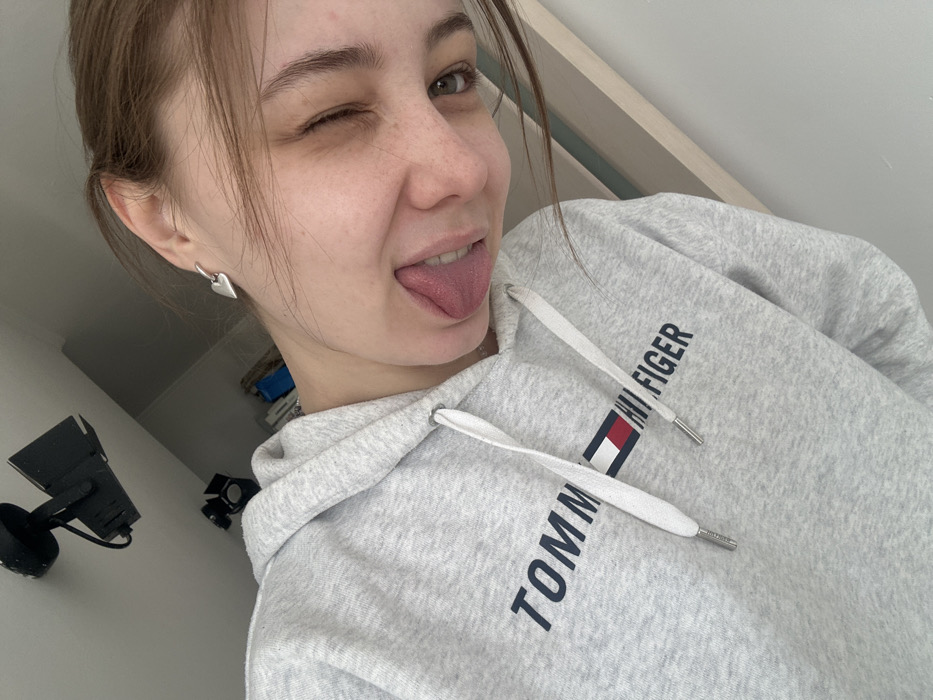 A young Caucasian female is depicted in a close-up, tilted selfie photograph taken indoors in Moscow, Russia. She is shown from the upper torso up, wearing a light grey Tommy Hilfiger hooded sweatshirt with visible white drawstrings and silver aglets. Her left ear displays a silver heart-shaped earring. She has light brown hair, visible freckles on her nose and cheeks, and is winking with her right eye while extending her tongue playfully. In the background, two black track lighting fixtures are mounted on a light-colored ceiling. Below the lighting, an indistinct background includes what appears to be shelving or furniture with a blue object and other items, alongside a light-colored wall or door frame. The scene indicates a casual, domestic environment.