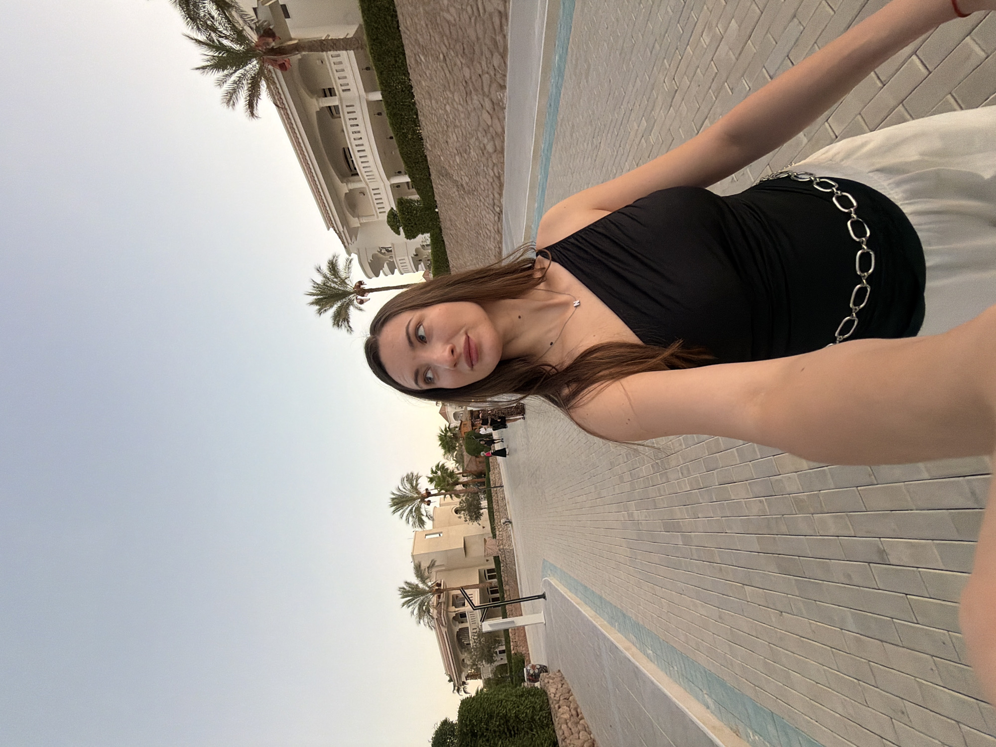 A woman with long brown hair is walking down a paved walkway in Sharm El Sheikh, Egypt. She is wearing a black one-shoulder top and white pants with a silver chain belt. In the background, there are palm trees, a large white building with balconies, and other people walking. The sky is clear and pale blue. The walkway is made of light-colored rectangular pavers arranged in a pattern, with a blue-lined border on the left. The overall scene suggests a warm, possibly evening, atmosphere in a resort-like environment.