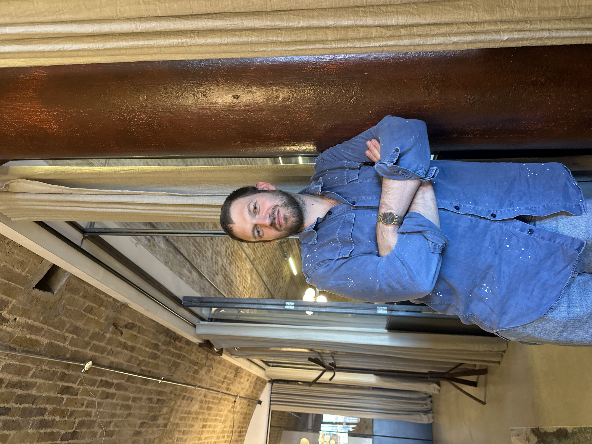 An indoor scene in a repurposed space, likely a railway arch in London, United Kingdom. A Caucasian male, approximately 30-40 years old, with short dark hair and a beard, is positioned centrally, leaning his left side against a dark reddish-brown cylindrical column. He faces the camera directly, displaying a slight smile. His arms are crossed over his chest. He wears a faded blue long-sleeved button-up shirt with scattered white paint splatters and light blue denim jeans. A gold-toned wristwatch with a rectangular face is visible on his left wrist.
The background features a vaulted brick ceiling with exposed metal conduit and a small white light fixture. To the man's left, a large glass partition with dark framing separates the immediate area from an adjacent room. Through this partition, a dark wooden coat rack is visible. Further in the background, additional light sources are present. To the man's right, a light-colored, textured curtain hangs adjacent to the column. The floor is a light, smooth surface.