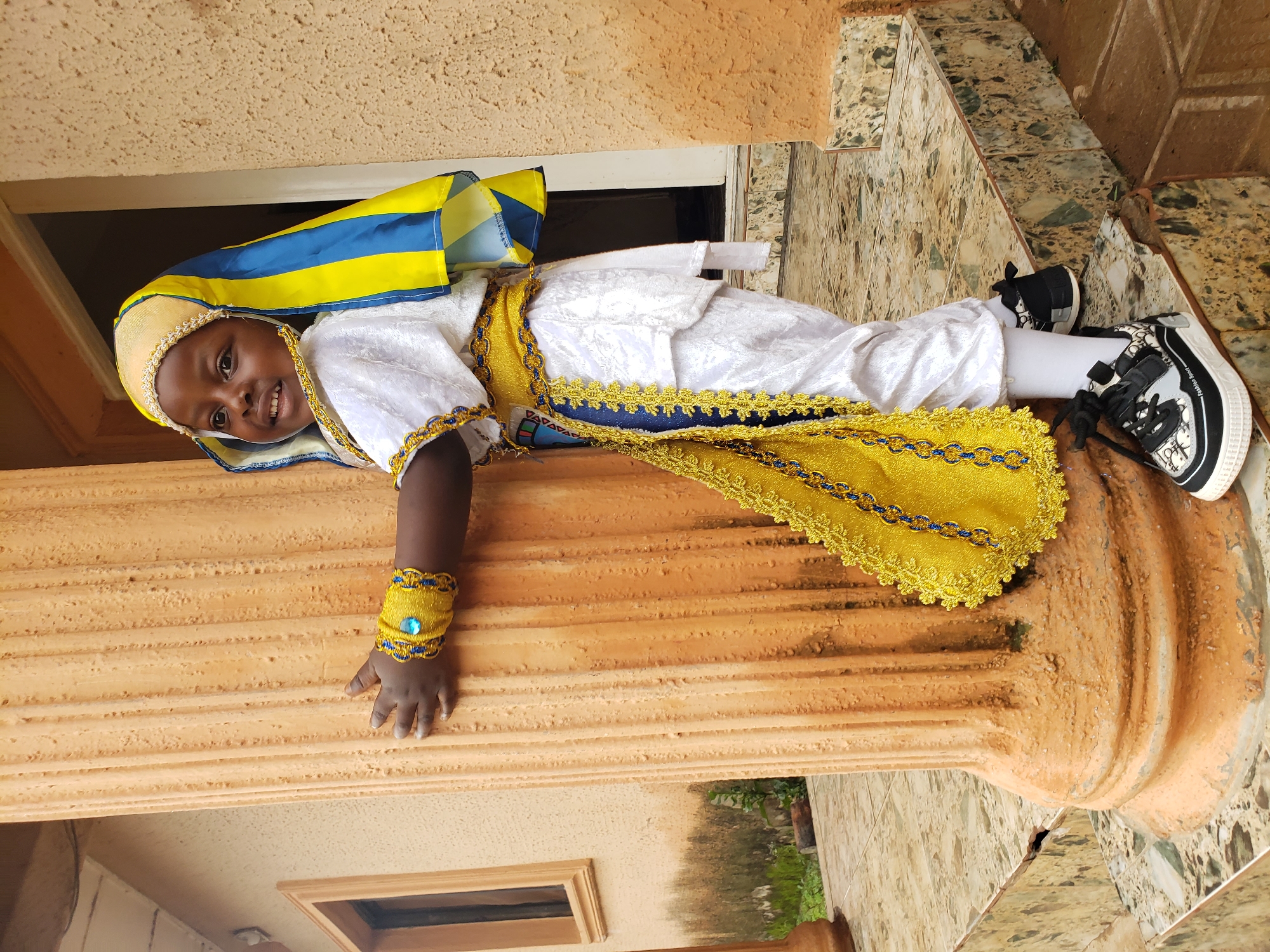 A young child, likely of Nigerian descent, stands on a tiled staircase, embracing a fluted, terracotta-colored column. The child is adorned in a white and yellow traditional outfit, complete with a head covering featuring blue and yellow stripes, and decorative gold and blue beaded bracelets on their wrists. The child's attire includes white pants and a white tunic with intricate gold trim and a yellow sash around the waist. The backdrop is a textured, light brown wall. The setting appears to be outdoors, with a window visible on the left side of the image and hints of greenery in the background, suggesting a residential area. The presence of the tiled staircase and the style of the architecture are consistent with urban environments in Nigeria, specifically the Sabon Tasha area of Kaduna State, known for its mix of traditional and modern housing. The child's stance and smile suggest a playful or celebratory mood.