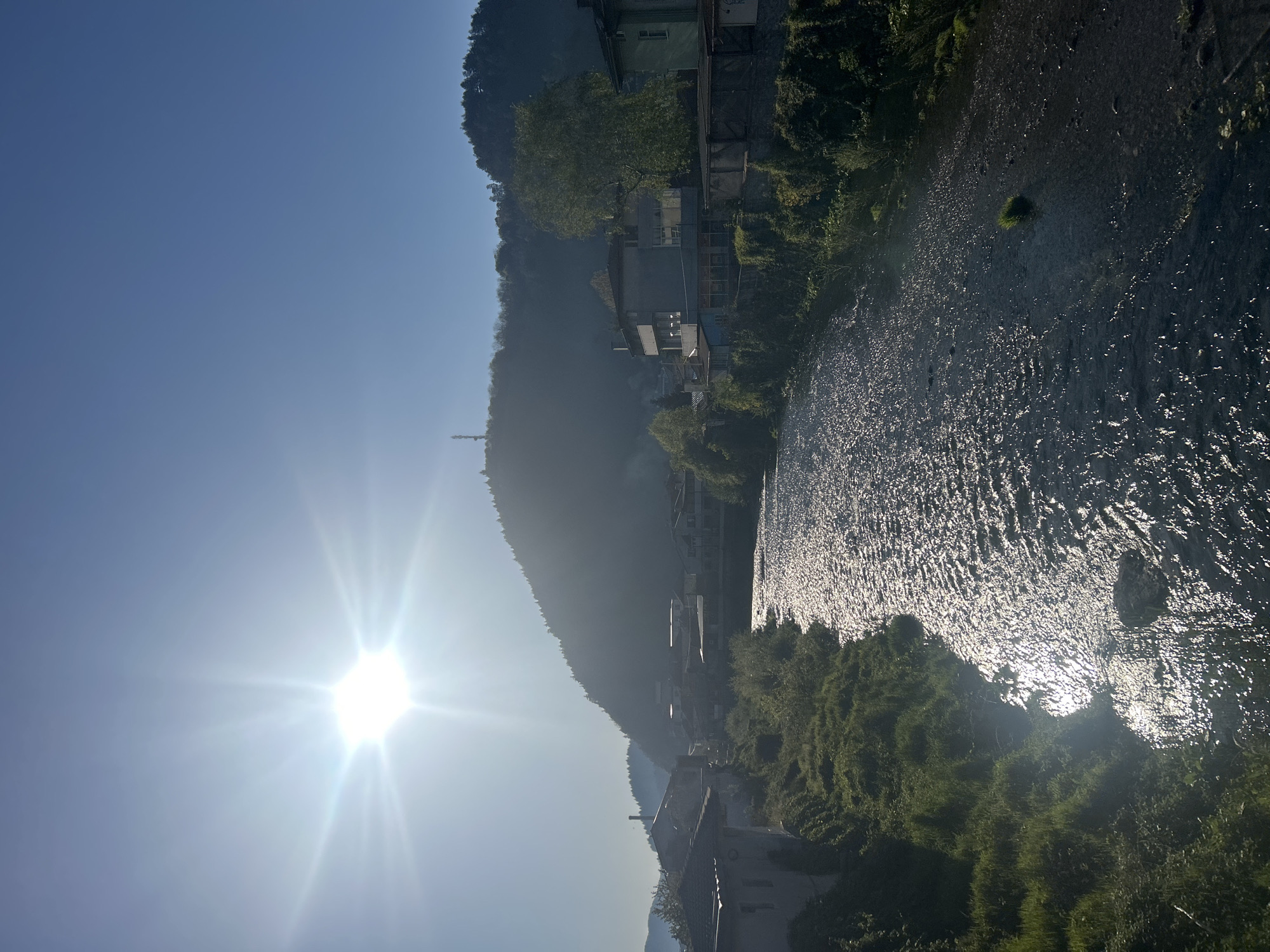 The image depicts a river in Zlatograd, Bulgaria, on a bright, sunny day. The sun is positioned on the left, casting a glare and multiple reflection points on the river's surface. The river bends to the right, bordered by trees and some houses. A tall, forested hill rises on the left side of the river, where additional houses are situated. The clear blue sky is visible in the upper left of the image.