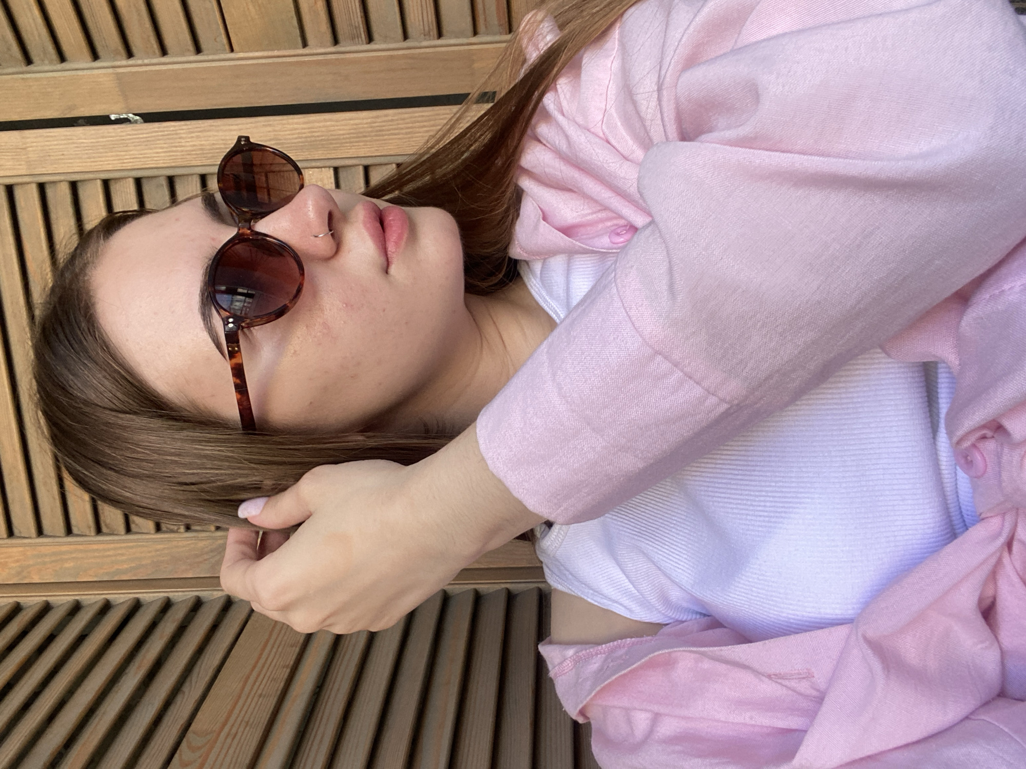 A young woman with long brown hair and sunglasses is pictured outdoors. She is wearing a pink unbuttoned shirt over a white tank top. She is touching her hair with her right hand. The background consists of wooden shutters. The location is not specified in the image.