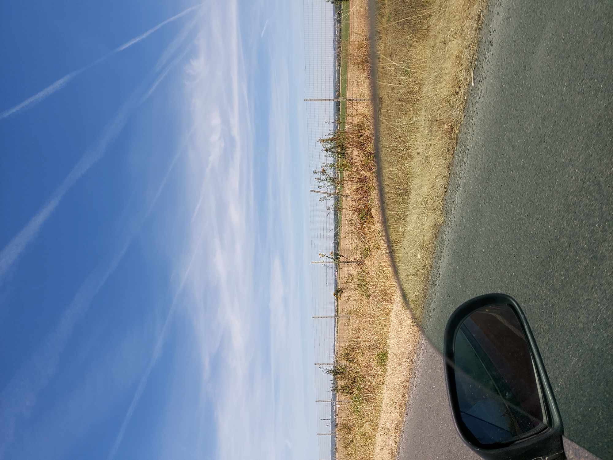 The image depicts a roadside view on a sunny day, likely taken from a moving vehicle. The foreground features the asphalt of a road with dry, yellow grass growing along its edge. A side-view mirror of a vehicle is visible on the left side of the frame, reflecting a dark, reddish-brown surface. In the middle ground, a wire mesh fence runs horizontally across the scene, separating a dry, grassy field from an area beyond. The field appears to be agricultural, with some sparse vegetation and a few distant structures, possibly wind turbines. The sky is a bright blue with numerous white contrails from airplanes streaking across it. The overall impression is one of a rural landscape under a clear sky. While the exact location in Avernes, France, is not explicitly indicated by any visible landmarks or text, the scene is consistent with a rural setting in that region.