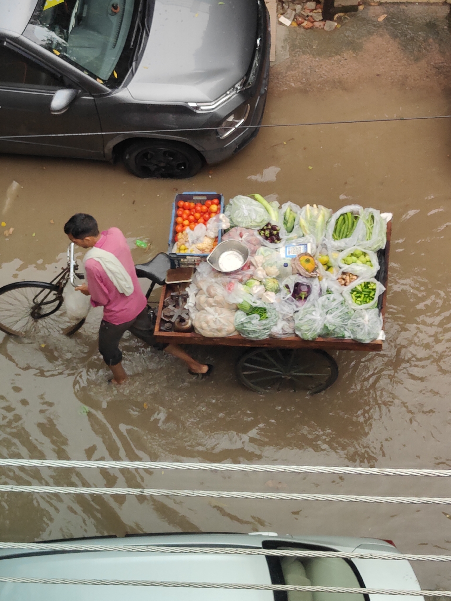 The image captures a street scene in Gurgaon, India, likely after heavy rainfall, as evidenced by the significant waterlogging. A man, dressed in a pink shirt and dark trousers, is pushing a vegetable cart. The cart is laden with a variety of produce, including tomatoes, cabbage, eggplant, chilies, and other vegetables, some of which are packaged in plastic. The man is wading through the water, which reaches his ankles. In the background, a dark-colored car is partially submerged in the water, with its front tire visible. The overall environment appears to be a residential or commercial street, with some debris visible on the sidewalk. The scene depicts a common sight in many Indian cities during monsoon season, where vendors continue their trade despite adverse weather conditions.