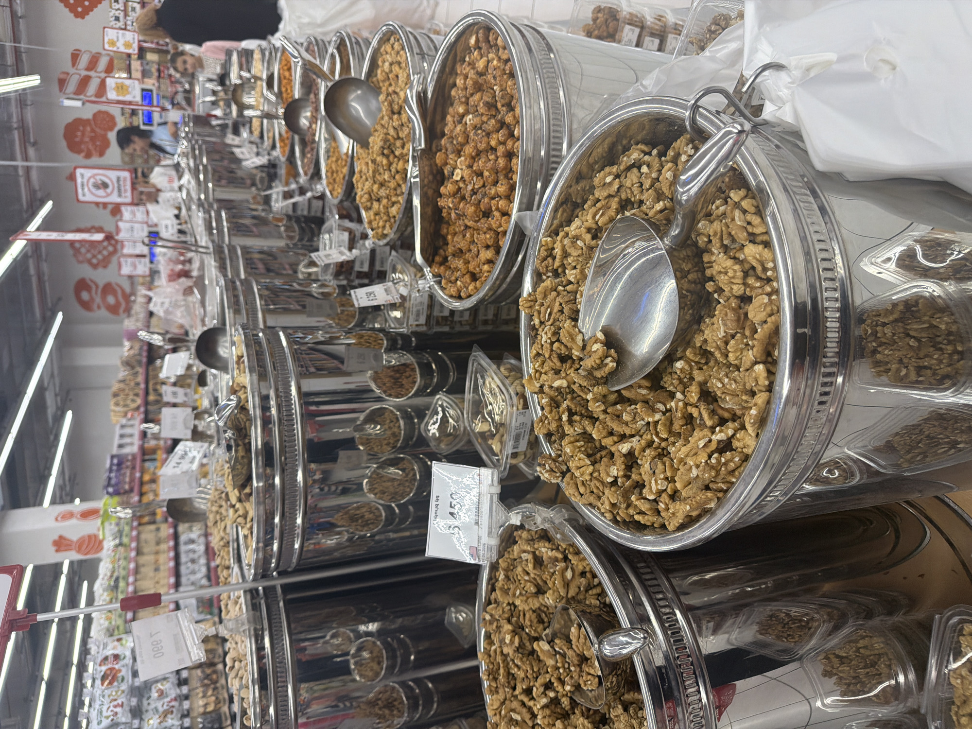 The image captures an aisle of a supermarket, likely in Yerevan, Armenia, showcasing a bulk-food section. Arranged are numerous silver-colored bins containing various types of nuts. Each bin has a serving scoop. The foreground features walnuts. Further down the aisle, other types of nuts and dried goods fill the bins. Customers are visible in the background, browsing the products. Supermarket shelving with various food items is seen in the distance along the aisle, and the store's ceiling lights illuminate the area. Signage and price tags are visible, adding to the supermarket atmosphere.