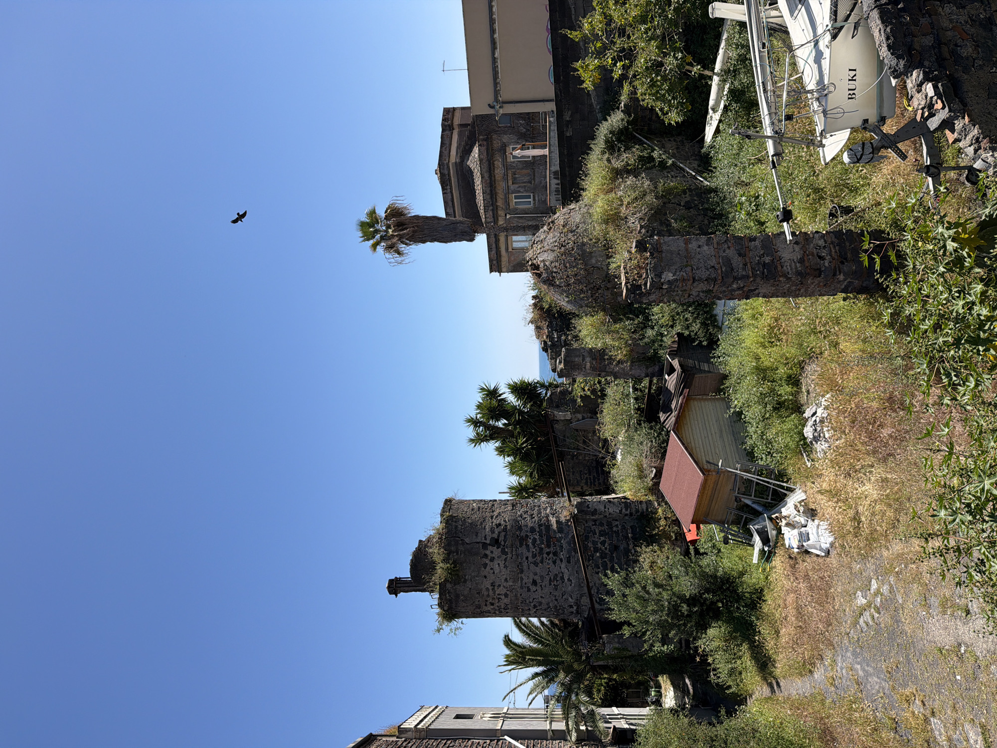 The image, rotated to an upright orientation, captures a sunlit, somewhat rugged coastal or hillside scene, likely in Catania, Italy, given the context. A clear, brilliant blue sky dominates the upper portion of the frame, with a single dark bird in mid-flight on the left.
The midground features a blend of natural growth and old structures. Prominently, a large, weathered stone tower or building remains, heavily overgrown with vegetation. To its right, a tall palm tree stands. Further back, a more complete building with dark stone walls, multiple windows, and a balcony is visible. On this balcony, a person dressed in light clothing stands, facing left.
The right side of the image reveals a white sailboat, marked with the text "BUKI," seemingly stored on land amidst overgrown grass and scattered debris. Its mast and rigging are visible. Below this, a rustic area features a small wooden shed with a red roof and more dry and green vegetation, suggesting a less maintained or abandoned plot of land. Another robust, dark stone structure, possibly a wall or ruin, is present on the far left, partially obscured by green foliage. Glimpses of the sea can be seen in the distance, a deep blue hue beneath the horizon. The bright light and clear sky indicate a sunny day, likely morning or early afternoon.