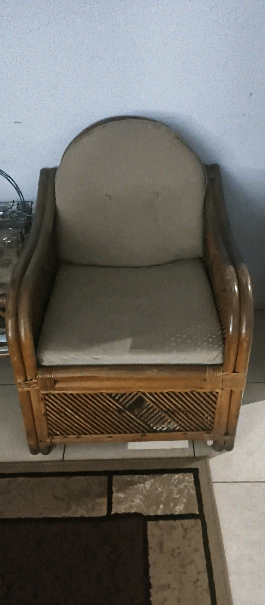 A light brown, woven rattan armchair with light beige cushions is centrally positioned in a room. The armchair has rounded arms and a curved back. The base of the chair features a diamond-shaped woven pattern. The armchair rests on a floor covered by a rug with a dark brown center and lighter brown and gray rectangular accents. The background consists of a plain, light-colored wall. To the left of the armchair, a portion of a dark metal object, possibly a side table or lamp base, is visible. The scene appears to be an interior domestic setting.