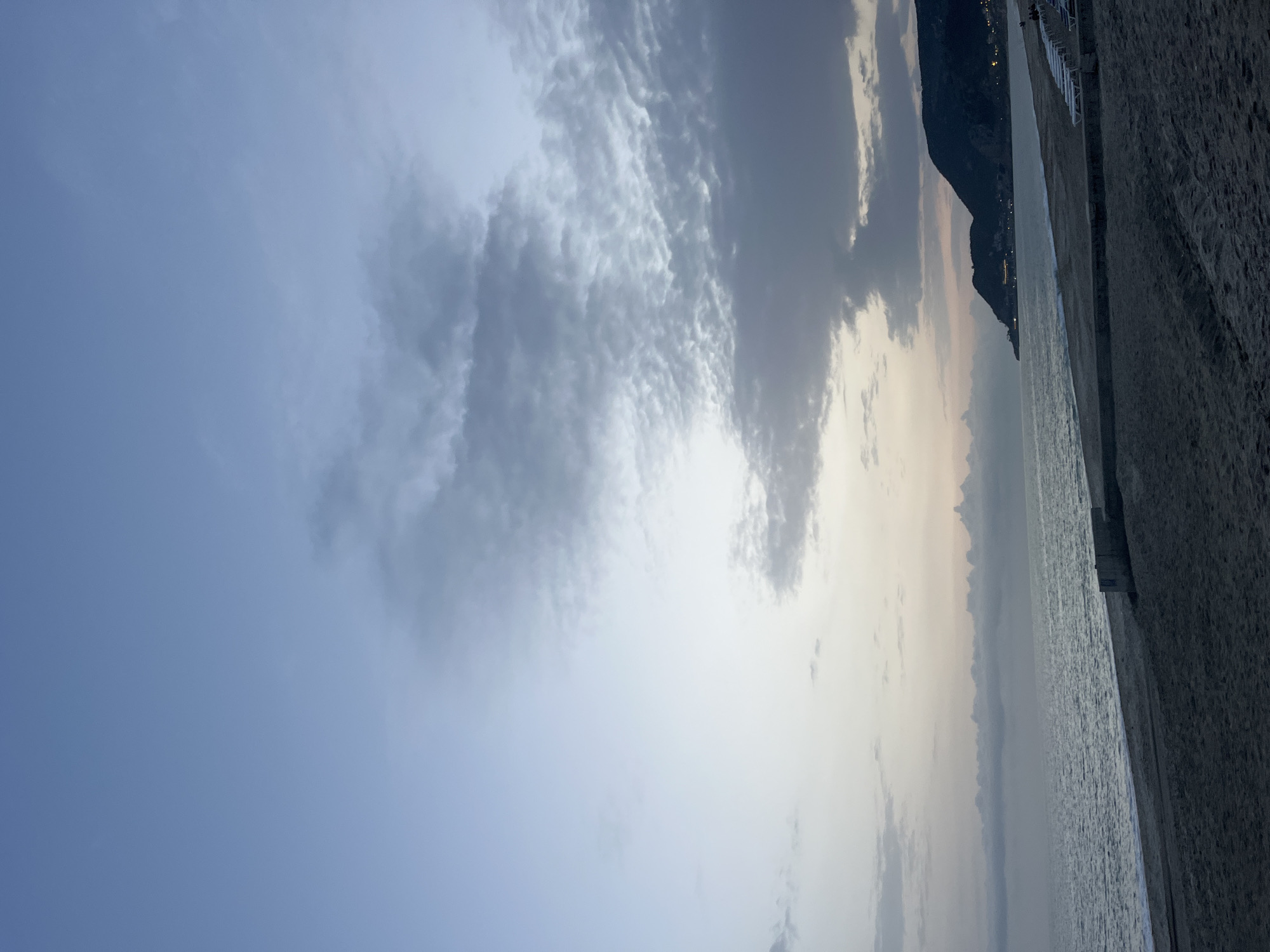 The image depicts a dusky seascape with a beach in Alanya, Turkey. The sky is predominantly blue with patches of cumulus clouds, transitioning to a muted sunset hue near the horizon. A mountain is visible along the coast, with some lights suggesting a populated area. The sea is calm and reflective. The beach is composed of sand. The image is oriented in landscape format.