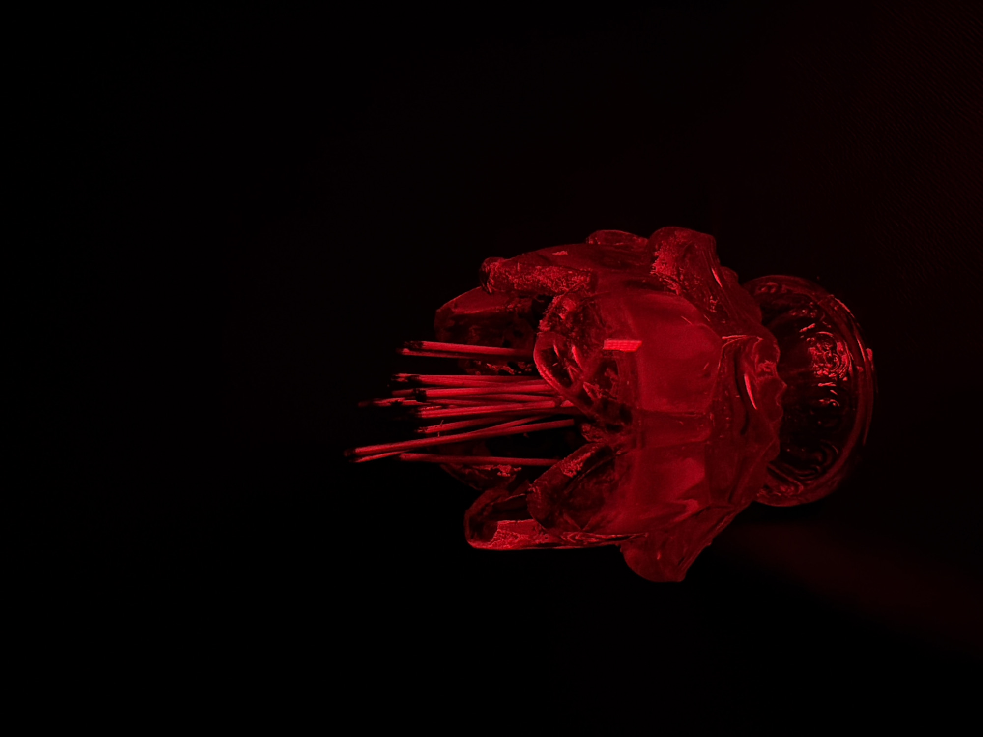 A red incense holder, shaped like a lotus flower, sits on a dark surface. Several unlit incense sticks are placed upright in the holder, which appears to be made of glass. The scene is illuminated by a red light, casting a faint shadow to the left of the holder. The background is entirely black, creating a stark contrast with the red object. There are no people present in the image.