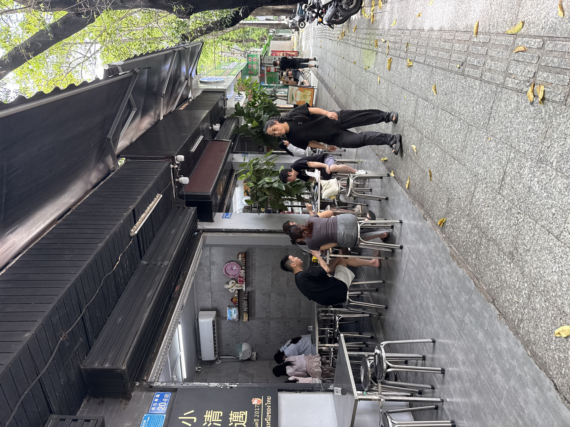 The image depicts a street scene outside a restaurant in Huizhou, China. In the foreground, a woman in black clothing walks on a sidewalk. The sidewalk is paved with granite tiles and is littered with fallen leaves. To the left of the sidewalk is the outdoor seating area of a restaurant, with tables and metal stools. Several patrons are seated at the tables, eating and drinking. The restaurant itself appears to be open, with an awning extending over the seating area. In the background, there are more buildings and trees. The overall impression is of a busy urban environment.