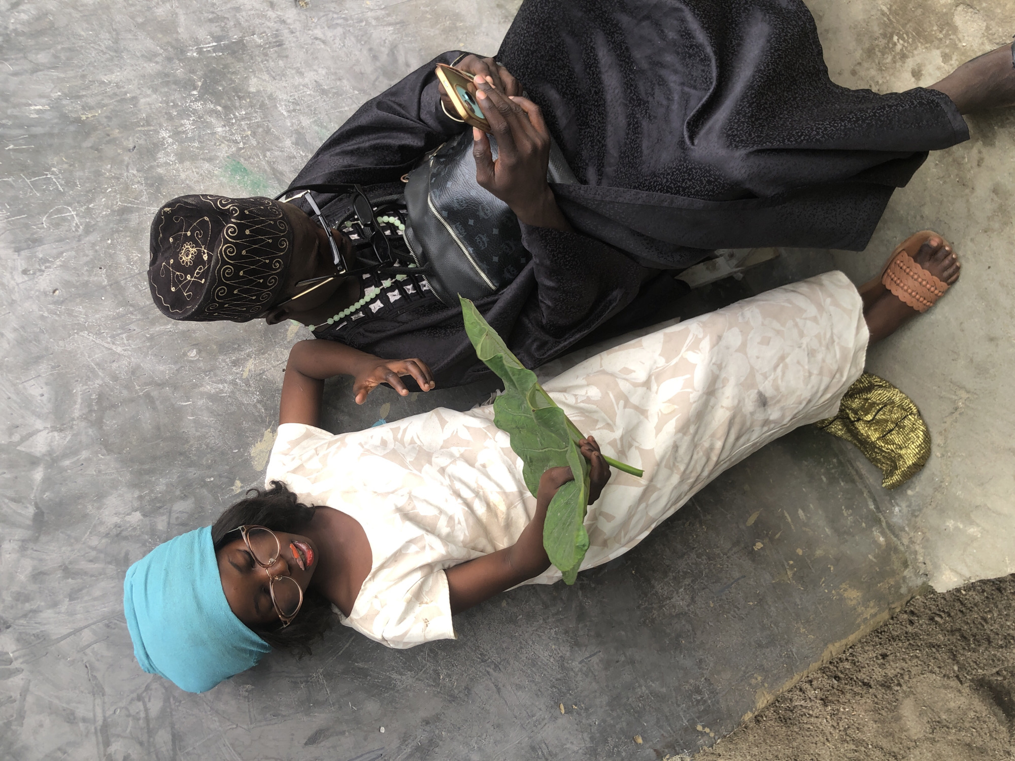 Two individuals are situated leaning against a textured concrete wall, with one seated and the other reclining. The seated individual, on the right, wears a black traditional robe and a black cap adorned with gold embroidery. They are looking down at a mobile phone held in their hands and are wearing glasses. A black bag with a distinctive pattern rests on their lap. The reclining individual, on the left, is a younger person wearing a light-colored dress and a vibrant blue headscarf. They have glasses with prominent frames and bright red lipstick. In their hands, they hold a large green leaf. The ground beneath them is a mix of dirt and concrete. A small, crumpled piece of gold fabric is visible near their feet. The overall setting appears to be an outdoor urban environment, possibly in Ado, Nigeria, given the style of dress and architectural context of the wall.