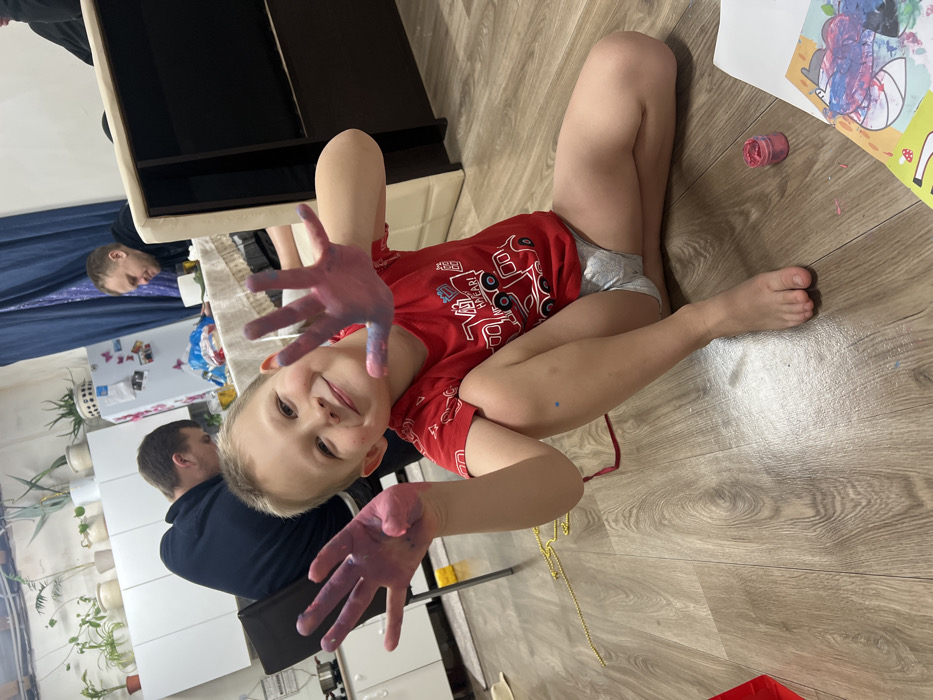 A young boy with paint-covered hands lies on a wooden floor, smiling at the camera. He wears a red shirt and light-colored underwear. An open jar of paint sits nearby. A drawing, also with paint, is on the floor next to him. Two adults are partially visible in the background near a white table and cabinets with plants. A cabinet and a blue curtain are behind them.