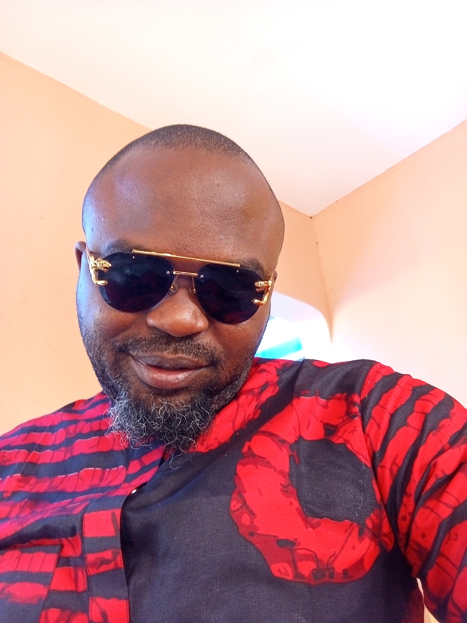 A medium shot depicts a man with dark skin, a shaved head, and a salt-and-pepper beard. He is wearing sunglasses with a gold frame and dark lenses. He is wearing a shirt with a black base and red horizontal stripes with a large red circular design on the front. The background is a pale peach-colored wall. The image was taken in Ankpa, Nigeria.