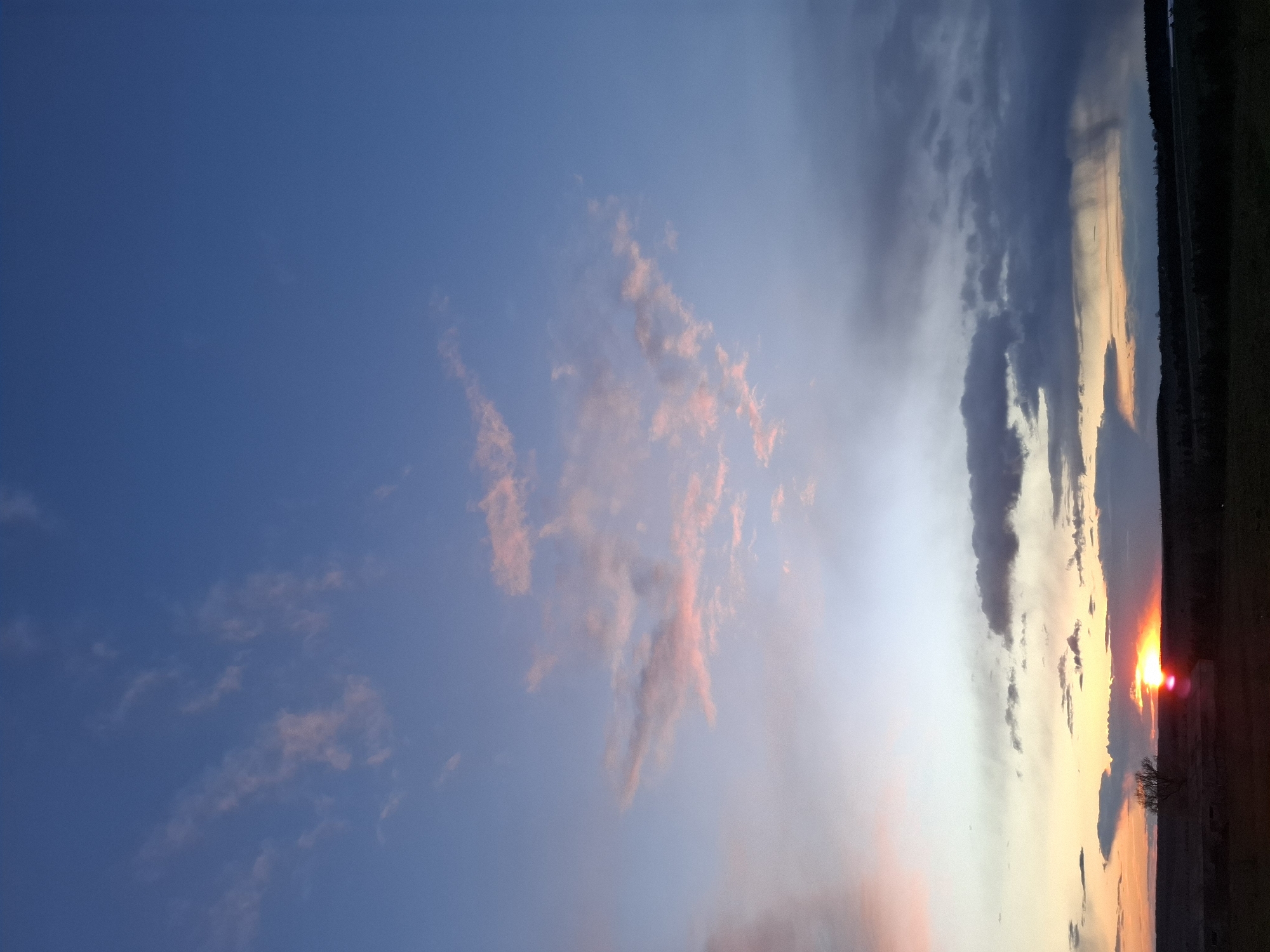 The image displays a serene sunset over a landscape, with the sky as the dominant feature. The scene is oriented vertically, but when viewed horizontally (rotated 90 degrees counter-clockwise), the sun is setting on the right horizon.
The sky transitions from a clear, deep blue in the upper left to lighter blues, purples, and grays towards the right, where the sun is located. Scattered across the mid-section of the sky are delicate, wispy clouds, tinged with soft pink and orange, catching the last light of the day. Towards the right side, closer to the horizon, darker, heavier clouds in shades of grey and blue are present.
The reddish-orange sun is visible on the far right, just above the horizon, casting a bright, warm glow that illuminates the lower clouds in brilliant oranges and yellows. Below the sun and extending across the horizon is a dark, silhouetted strip of land, suggesting distant trees or structures, characteristic of a rural setting. This landscape is consistent with the provided location context of Khlevnoye, Russia. The time of day is clearly sunset, creating a tranquil and visually dramatic atmosphere. No people, specific objects, or visible text are present in the image.