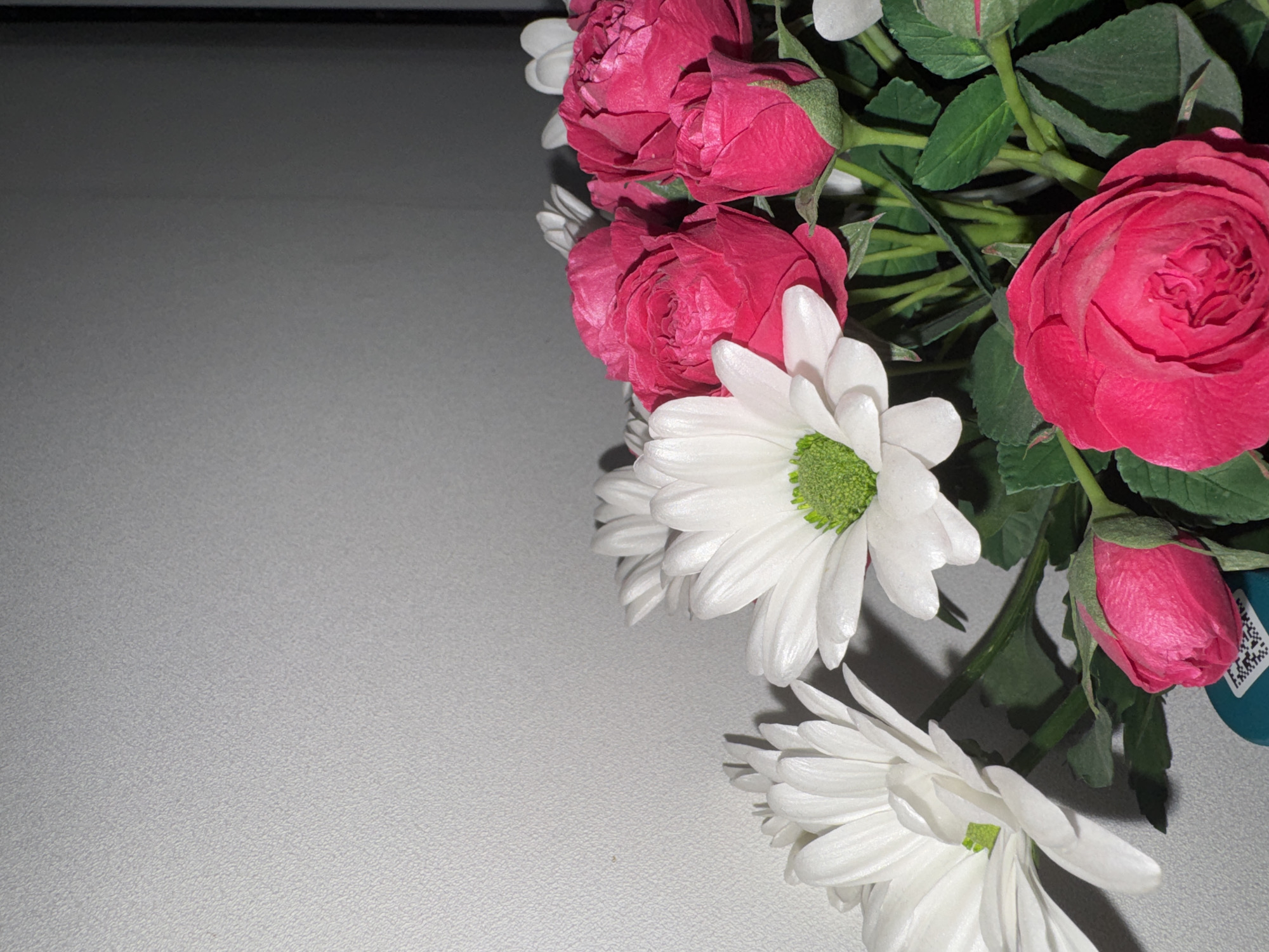 A close-up, still life photograph, potentially taken indoors in Chaikovsky, Russia, features a bouquet of flowers arranged against a light grey background. The flowers, primarily vibrant pink roses and white daisies, are clustered on the right side of the frame.
The bouquet includes several pink roses, appearing in various stages from tightly furled buds to fully bloomed flowers, showcasing a rich magenta or fuchsia color. Interspersed among the roses are white daisies, each characterized by numerous delicate white petals surrounding a distinct green or yellowish-green central disc. Green stems and leaves provide a natural backdrop to the blossoms.
The left and bottom portions of the image are dominated by a flat, light grey surface that exhibits a subtle, finely textured appearance, possibly a tabletop or counter. Soft shadows from the flowers are cast onto this surface, suggesting a diffused light source from the upper left. A darker grey area is visible along the top edge and behind some of the flowers, indicating a deeper background or a wall.
A small, dark teal object, possibly part of a vase or a decorative tag, is partially visible behind a rose bud on the lower right. This object features a small white square containing an intricate, pixelated pattern, likely an illegible QR code or barcode. No explicit text is discernible. No specific activity is depicted, making it a static representation of a floral arrangement.