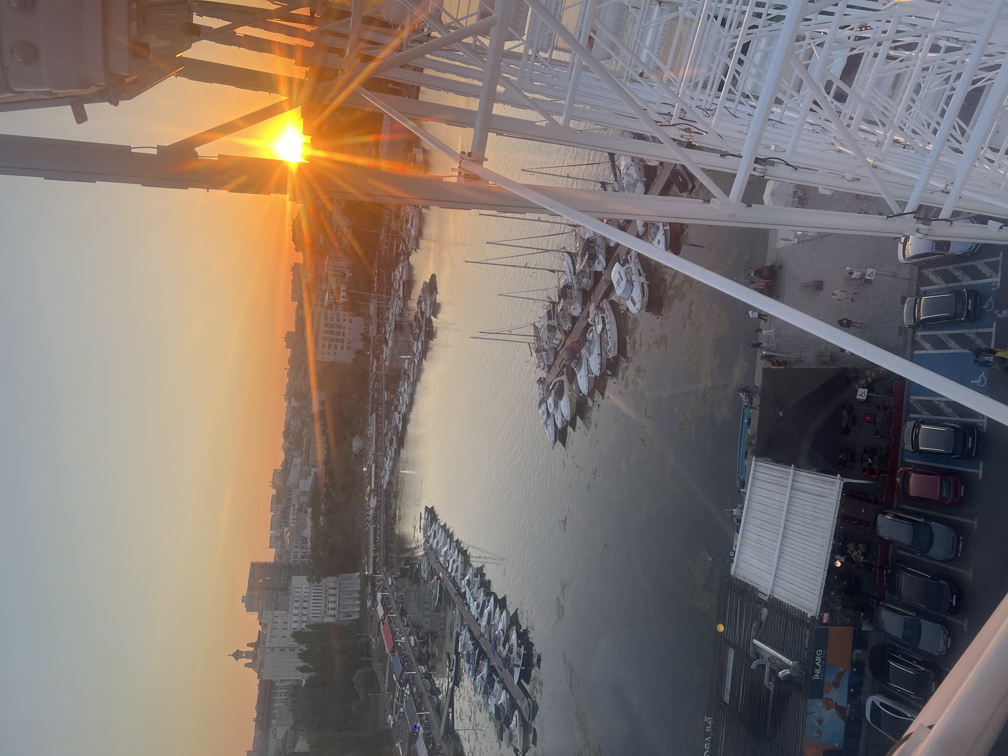 The image captures a sunset over the city of Constanța, Romania, as viewed from a Ferris wheel. In the foreground, the white lattice structure of the Ferris wheel dominates the right side of the frame. Below, a marina is filled with numerous docked boats of various sizes, including speedboats and sailboats. The water of the marina reflects the warm hues of the setting sun.
Beyond the marina, the city skyline is visible, with buildings nestled on a hill. The sun, low on the horizon, is a bright orb with prominent sunburst rays, creating a vibrant orange and yellow glow that illuminates the sky. The sky transitions from a soft orange near the sun to a pale blue at the top of the frame.
On the left side of the marina, there are waterfront structures that appear to be restaurants or cafes, with tables and umbrellas visible. A row of parked cars is also present on the left, adjacent to a building with signage that includes the word "INLARG". Several people are seen walking along the waterfront and near the parked cars. The overall scene depicts a serene evening at a coastal city's harbor, with the dramatic backdrop of a sunset.