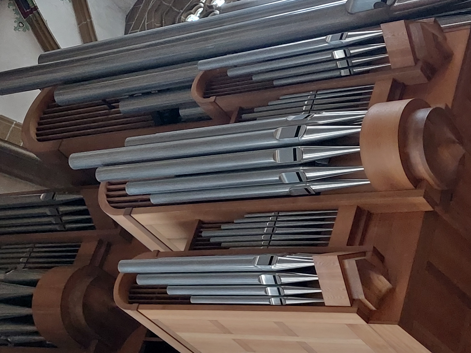 The image shows a close-up view of a section of a pipe organ, likely located within a church or cathedral in Erfurt, Germany. The organ is constructed primarily of wood and metal pipes of varying sizes, with a prominent arrangement of polished metal pipes in the foreground. These pipes are arranged in tiered rows, with the largest pipes on the left and progressively smaller pipes to the right. The wooden casing of the organ features decorative elements and a warm, polished finish. The background reveals more of the organ's structure, including darker, larger pipes and architectural details of the building, such as arched ceilings and stained glass windows, though these are out of focus. The lighting suggests an interior setting, possibly with ambient light filtering in from windows. There are no people visible in the frame.