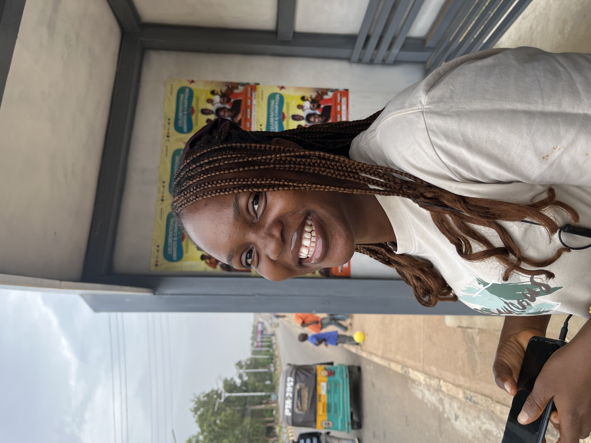 A woman with brown braids is smiling at the camera. She is wearing a beige t-shirt with a muted green and blue design. She's holding a black phone in her right hand. She is standing at a bus stop with advertisements behind her. Outside the bus stop, there is a blue and yellow tuk-tuk traveling on the road. There are also people, including a child kicking a yellow ball on the sidewalk in the background. The license plate of the tuk-tuk is PTA-2057. The location appears to be the city of Bar Jirgi Summa in Nigeria, given the context of the information provided.