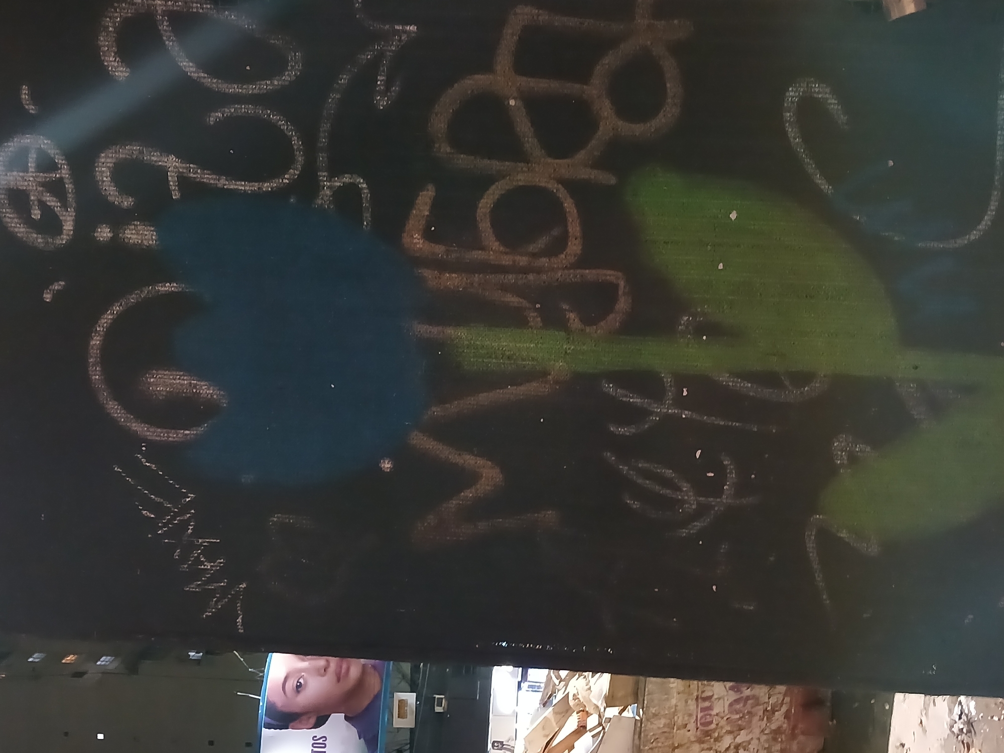 The image displays a vertical section of a dark wall or surface, heavily adorned with white and light-colored graffiti in a cursive and calligraphic style, characteristic of urban environments in Buenos Aires, Argentina. The graffiti features numerous swirling lines, abstract shapes, and partially legible characters. Among these, numbers like "2," "8," and "9" are discernible, as are letters such as "c," "L," "g," "z," "e," "r," "s," "d," "y," and "p." Two large, indistinct spray-painted shapes in blue and green are also present, overlapping parts of the white graffiti. A strong light source creates a bright glare across the upper left and central portions of the wall, illuminating the textures of the graffiti.
Below this graffiti-covered surface, a large poster or screen is visible, featuring a close-up of a young woman or teenager. She has dark hair, dark eyes, and a neutral expression, looking slightly upwards. She is wearing a white and purple garment. Partial text is visible on the poster: the letters "TOS" can be seen below her chin, and the word "SOL" is visible on the bottom left corner of her shirt. The background behind the poster shows indistinct urban elements, possibly buildings or street infrastructure, with some torn papers and debris at the very bottom, suggesting a street-level environment. The image is a static capture of an urban scene, with no specific activity or event taking place.