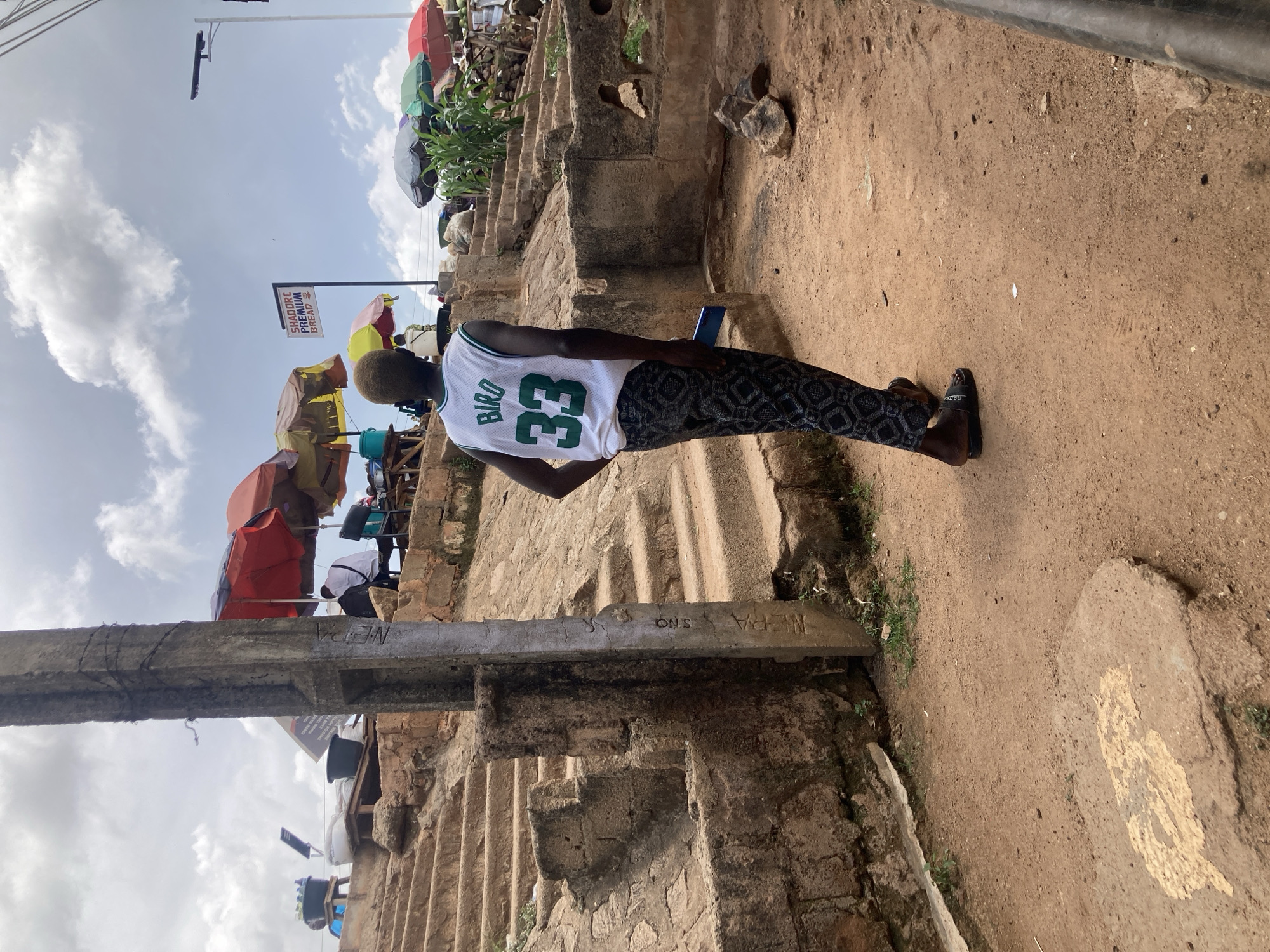 The image depicts an outdoor scene in Gwong, Nigeria, likely a marketplace area with a sandy ground and stone steps. In the foreground, a person is seen from behind, wearing a white basketball jersey with "BIRD" and the number "33" printed on it, and patterned dark pants. They are holding a blue object, possibly a phone, in their right hand. Behind this person, a concrete pillar stands to the left, with some writing on it. To the right, the ground slopes upwards with stone steps and a retaining wall.
Further in the background, various stalls with colorful umbrellas are set up, suggesting commercial activity. Some people are visible sitting or standing at these stalls. A sign that reads "SHADDORC PREMIUM BREAD" is attached to a pole. The sky is partly cloudy. The overall impression is of a bustling, informal commercial environment.