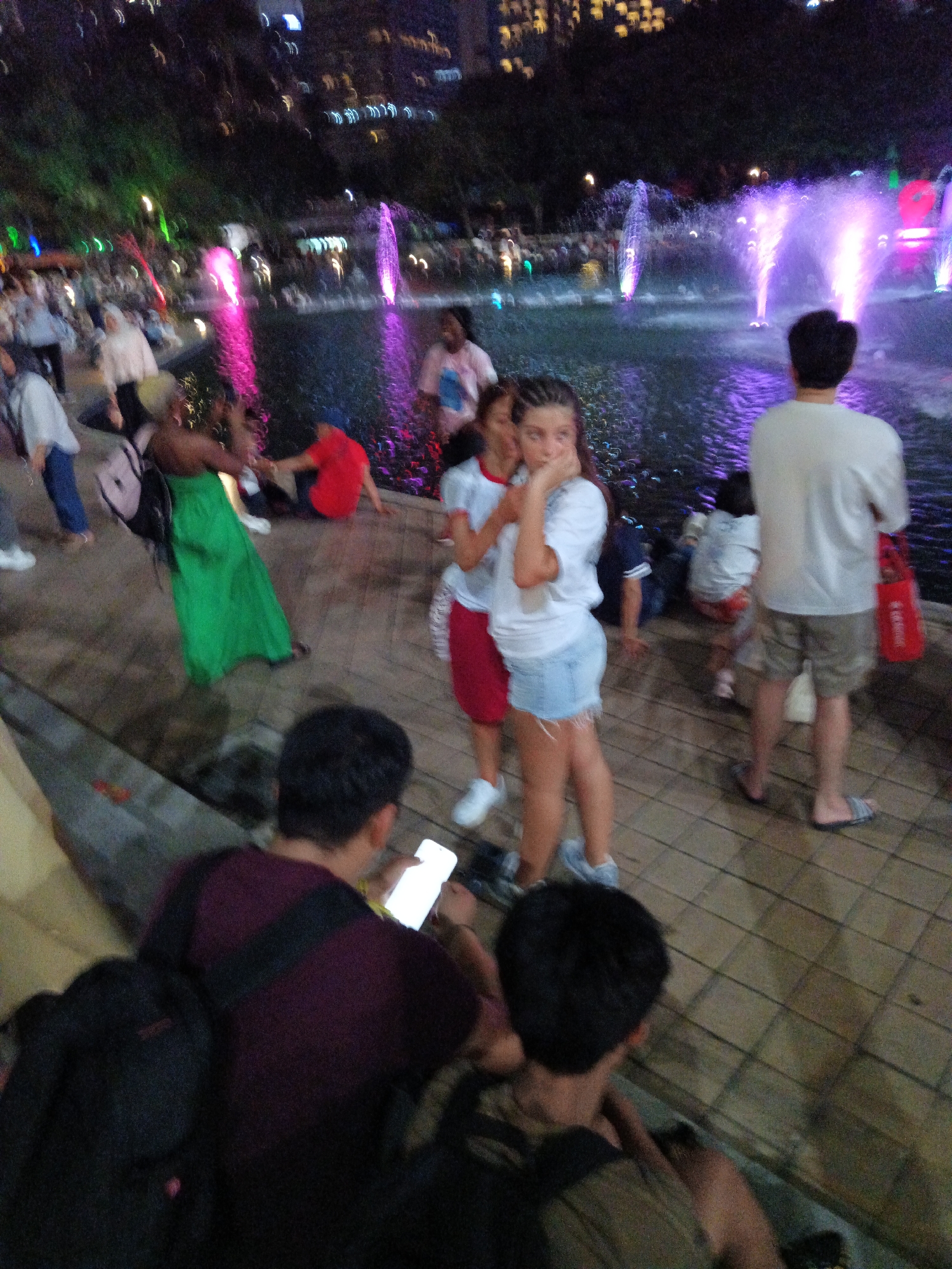 The image depicts an outdoor scene at night, likely in a public park or recreational area. In the background, illuminated fountains spray water with purple and pink lights, suggesting a decorative water feature. City lights and buildings are visible in the far background, indicating an urban setting.
In the foreground and midground, several individuals are present. To the right, a man in a white t-shirt and khaki shorts stands facing away from the camera, looking towards the fountains. Nearby, a young woman in a white t-shirt and denim shorts is captured in profile, holding her hand near her face. Another woman in a white top and red skirt is partially visible behind her.
To the left, a person in a bright green dress is walking away from the camera, carrying a backpack. Several other individuals are scattered throughout the scene, some walking and others seated or crouching. One person in the lower left is looking at a phone. The ground is paved with square tiles. The overall atmosphere suggests a casual evening outing. The fountains and urban backdrop suggest this scene could be from KLCC Park in Kuala Lumpur, Malaysia, known for its decorative water features and proximity to city landmarks.