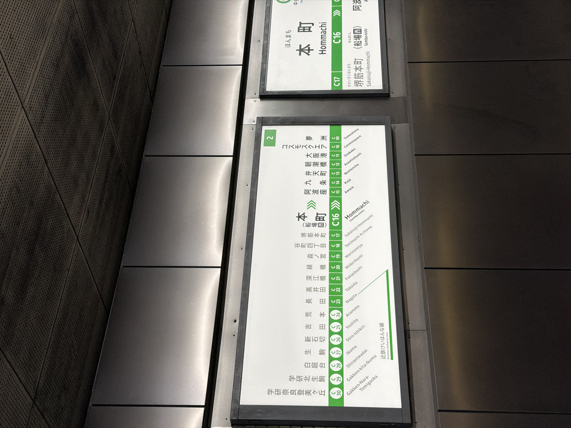 The image displays two illuminated information signs typically found in a Japanese subway station, viewed from a slightly tilted perspective. The signs are set against dark, textured wall panels on the left and smooth, metallic panels on the right, suggesting a modern underground environment.
The larger, main sign on the lower left presents a detailed list of stations along a subway line, identified by the 'C' prefix and numbers C09 to C30, indicating the Chuo Line. Station names are provided in both Japanese characters and Romanized script, including "Cosmosquare," "Osaka-ko," "Hommachi" (C16), "Sakaisuji-Hommachi" (C17), "Tanimachi 4-chome," and "Nagata." It also indicates a connection to the "Kintetsu Keihanna Line." A green rectangular box with the number "2" is visible at the top left of this sign.
The smaller sign at the top right prominently features "本町" (Hommachi) with the station code "C16," indicating the current station. It also shows "堺筋本町" (Sakaisuji-Hommachi) with "C17" and "(船場図)" (Semba-nishi) below it, likely indicating the next station or a transfer point. Arrows on both signs suggest directions of travel along the line. No people or specific activities are visible, focusing solely on the station information. The lighting is artificial, illuminating the signs brightly against the darker background.