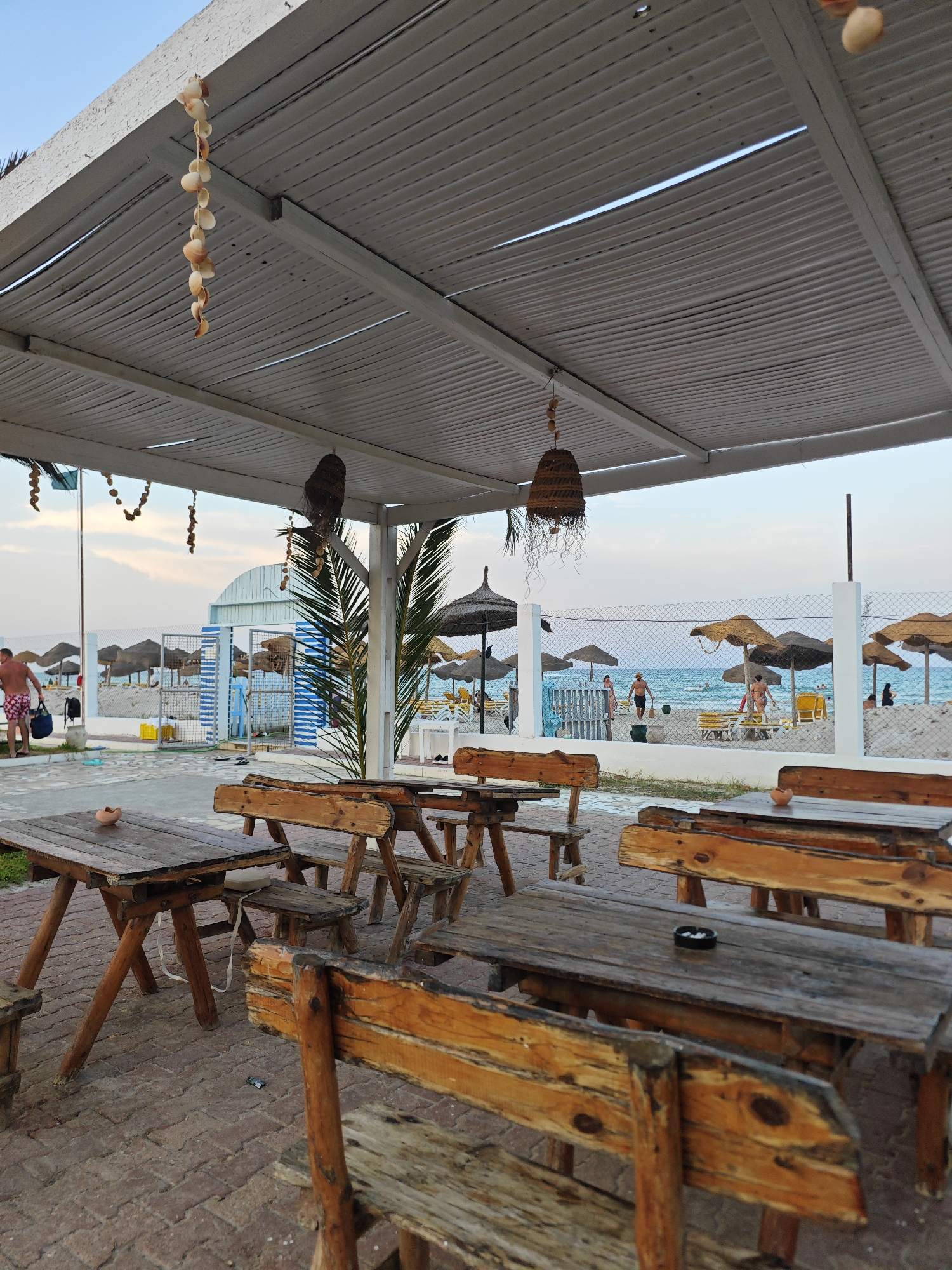 The image depicts an outdoor seating area with rustic wooden tables and benches situated on a brick-paved ground. In the background, a beach with umbrellas and people can be seen. The setting is likely a beachside cafe or restaurant in Mahdia, Tunisia, given the presence of Mediterranean beach elements like umbrellas and the general ambiance. The lighting suggests it could be late afternoon or early evening. Decorations of shells hang from the white canopy overhead, adding a coastal touch.