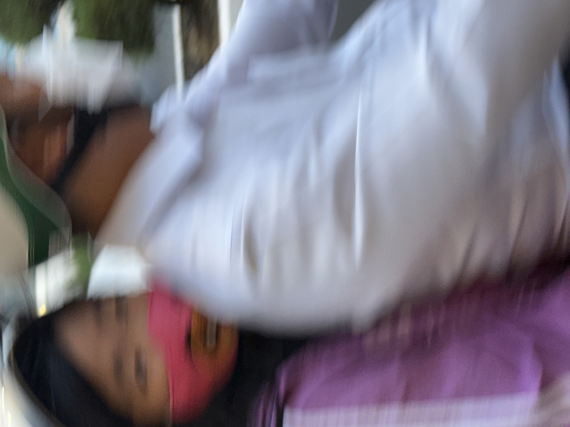 This media file shows a close-up, blurry view of two individuals, likely a child and an adult, outdoors. The child, on the left side of the frame, is wearing a pink mask that covers their nose and mouth, and has dark hair. Their face is partially visible, with eyes looking towards the camera. They are wearing dark clothing. The adult is positioned behind and above the child, wearing a white garment that appears to be a shirt or lab coat. The adult's face is not clearly visible due to the blurriness and their positioning.
The scene appears to be a daytime setting, possibly during warm weather, given the casual clothing and outdoor feel. There are hints of greenery in the background, suggesting a park or a natural environment. The overall impression is a candid, possibly informal, moment captured between the two individuals. The blurriness of the image suggests movement or a quick capture, making it difficult to ascertain specific details about their expressions or the exact activity. The colors in the image are muted, with prominent whites from the adult's clothing and a vibrant pink from the child's mask. There is also a section of purple fabric visible in the lower right corner, possibly part of the adult's attire or a nearby object. No text is visible in the frame. The context of Nha Trang, Vietnam, implies a tropical or sub-tropical environment.