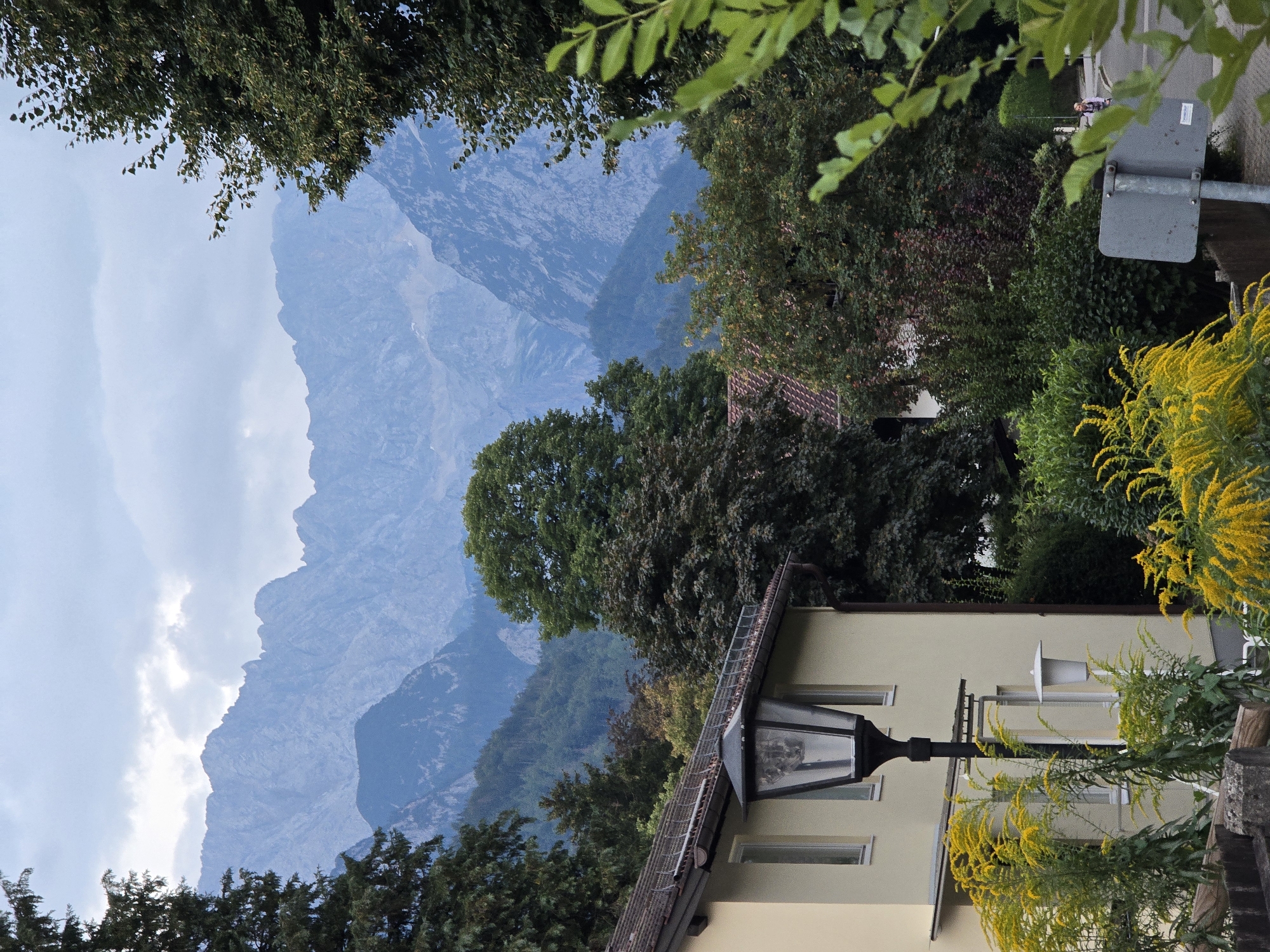The image shows a view of a street in Garmisch-Partenkirchen, Germany, with a backdrop of the Alps. In the foreground, to the left, is a portion of a light-colored building with windows and a black lantern post. Yellow flowering plants are visible in front of the building. To the right, a paved road curves away, and a person is seen riding a bicycle in the distance. A grey road sign is also present on the right side of the road. The midground is filled with lush green trees and foliage, with glimpses of red-tiled roofs of houses. The background features a dramatic, misty panorama of the rugged, blue-hued mountain range of the Alps. The sky is overcast with soft clouds.