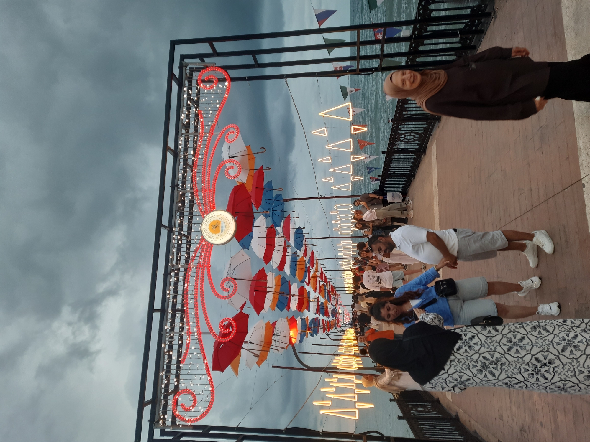 This image captures a festive scene on a pier in Atakum, Turkey. The pier is adorned with numerous colorful umbrellas suspended overhead, creating a vibrant canopy. String lights in the shape of triangles and circles illuminate the pathway, adding to the celebratory atmosphere. A decorative archway, embellished with red swirls and white lights, marks the entrance to the decorated section of the pier. Flags of various nations are also visible along the railing. A diverse group of people are strolling along the pier, engaged in conversation or simply enjoying the ambiance. Some are dressed casually, while others wear traditional attire. The sea is visible on both sides of the pier, with choppy water under a cloudy sky, suggesting an overcast day. The overall impression is one of a lively public gathering space decorated for an event.