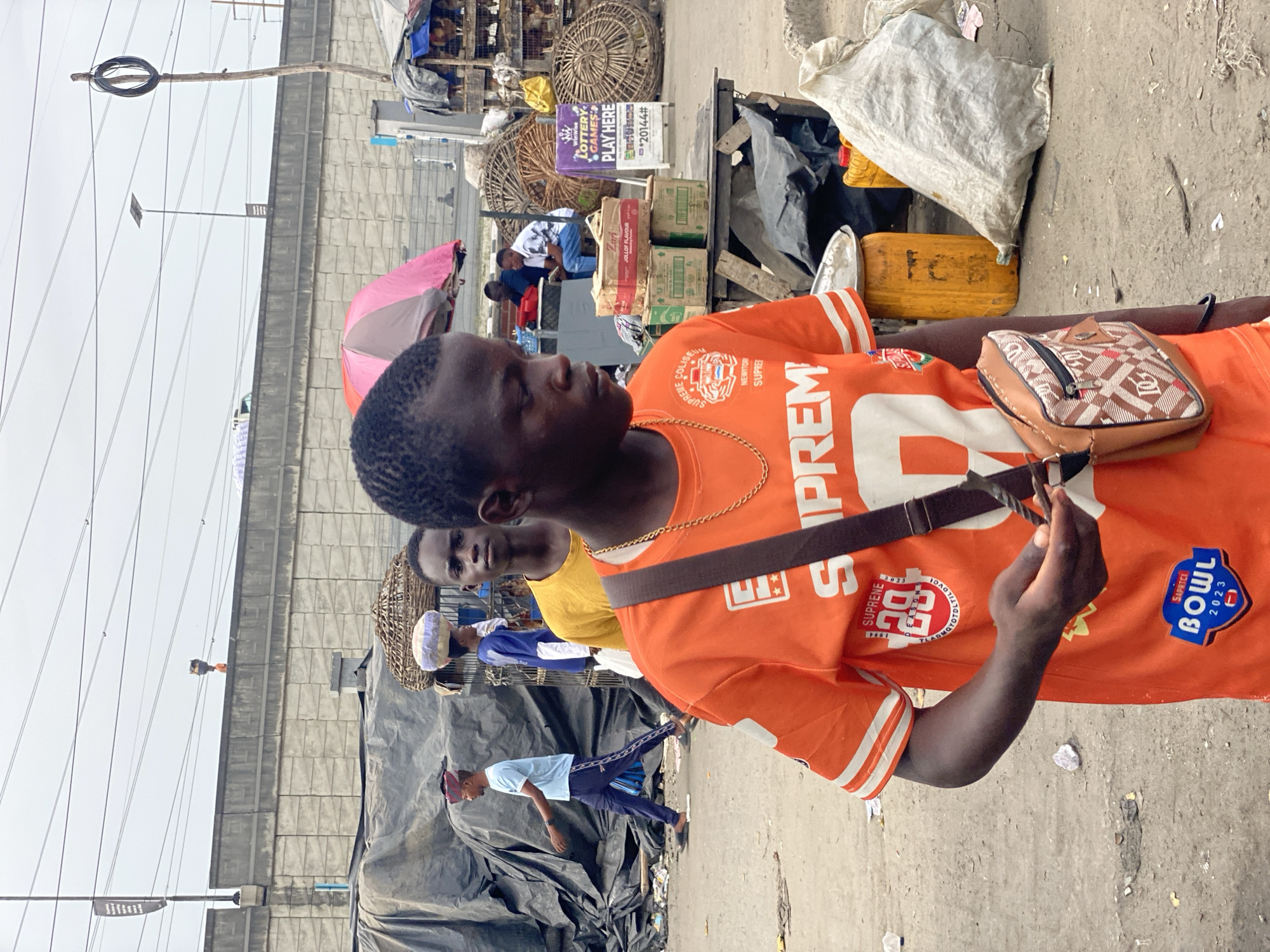 The image shows a street scene in Lagos, Nigeria. A young man wearing an orange Supreme jersey and a shoulder bag is standing and holding something in his hand. Behind him, there are other people walking and standing. Stalls with goods for sale, including what appears to be poultry in cages, are visible. There is an advertisement for lottery games. A pink umbrella is also visible in the background. The street appears to be cluttered with trash. Overhead power lines are present. The buildings are constructed of concrete blocks.
