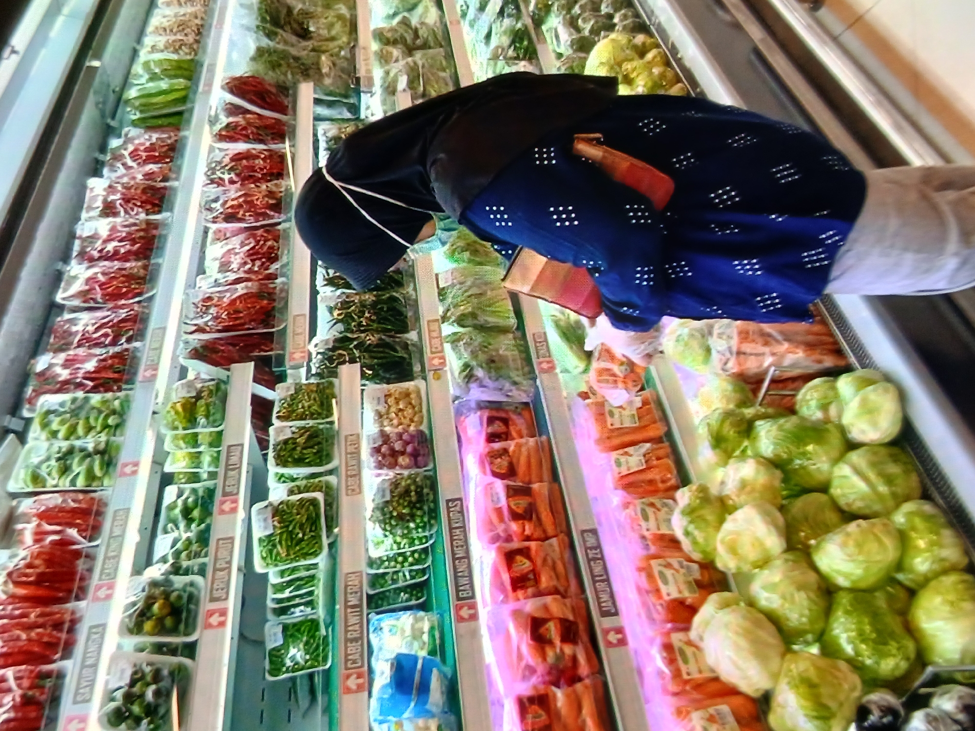 In a brightly lit supermarket aisle in Setia Bakti, Indonesia, a woman wearing a black hijab and a face mask browses a display of produce. She is dressed in a dark blue top adorned with white polka dots and light-colored pants. In her gloved hands, she holds an orange book or folder. The shelves are stocked with an array of fresh vegetables, packaged in transparent plastic. To her left, rows of lettuce heads are piled high, while further down the aisle, packaged carrots and other root vegetables are visible. Labels with Indonesian text indicate the types of produce, such as "SAYUR NANGKA," "JERUK PURUT," and "CABE RAWIT MERAH." The overall environment suggests a typical grocery shopping experience.