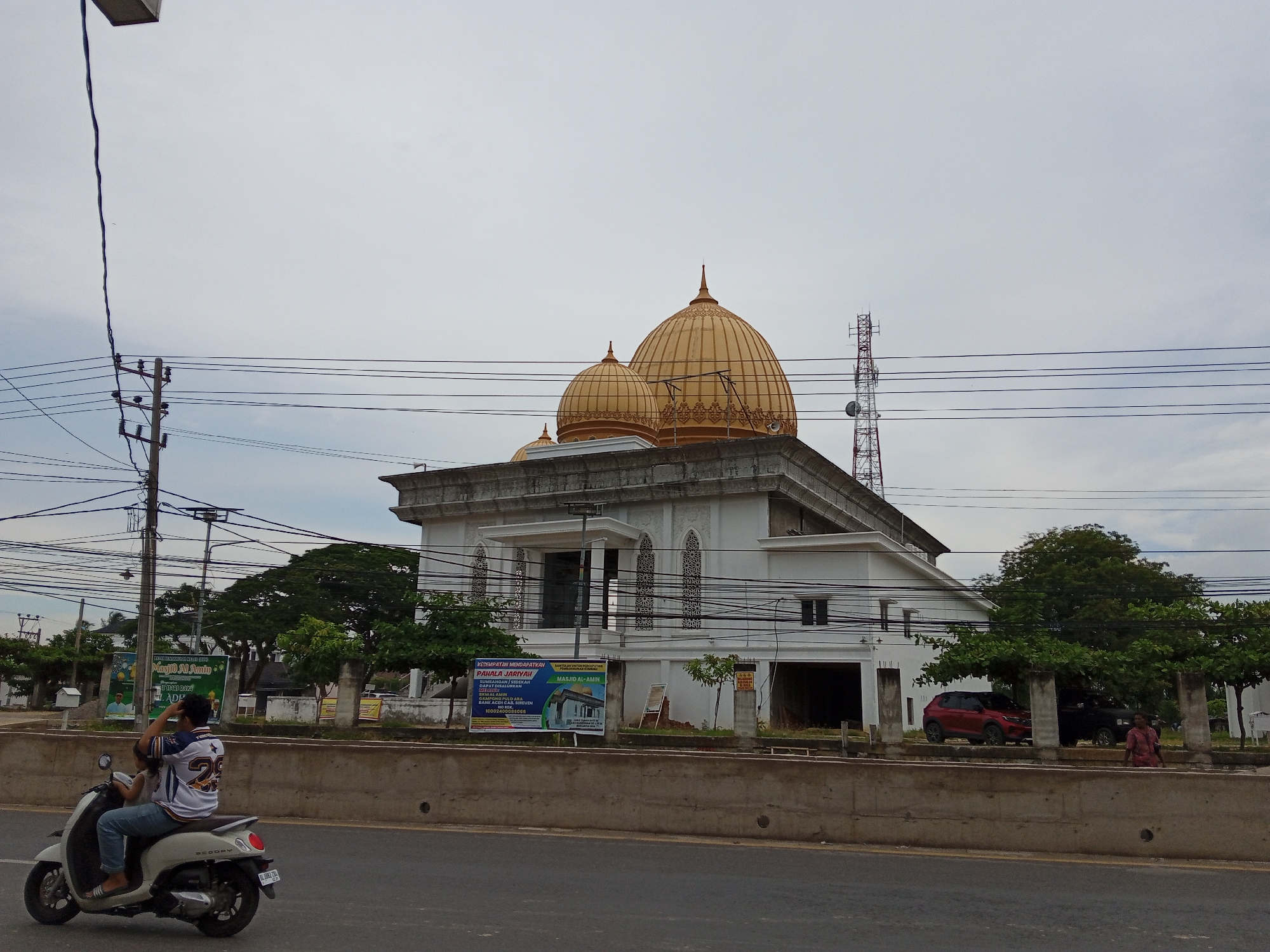 The image shows a mosque with two prominent golden domes in Puloara, Indonesia. The mosque is a white building with intricate decorative elements around the windows and a large entrance. In the foreground, a person is riding a white scooter on a paved road. The scooter is carrying a child as a passenger. The person on the scooter is wearing a white and blue shirt with the number 29 on the back. To the left of the mosque, there is a telephone pole with numerous wires. Behind the mosque, there are several trees and a red car parked in front of the building. A red and white telecommunications tower is visible in the distance. The sky is overcast and a concrete barrier runs along the side of the road in front of the mosque.