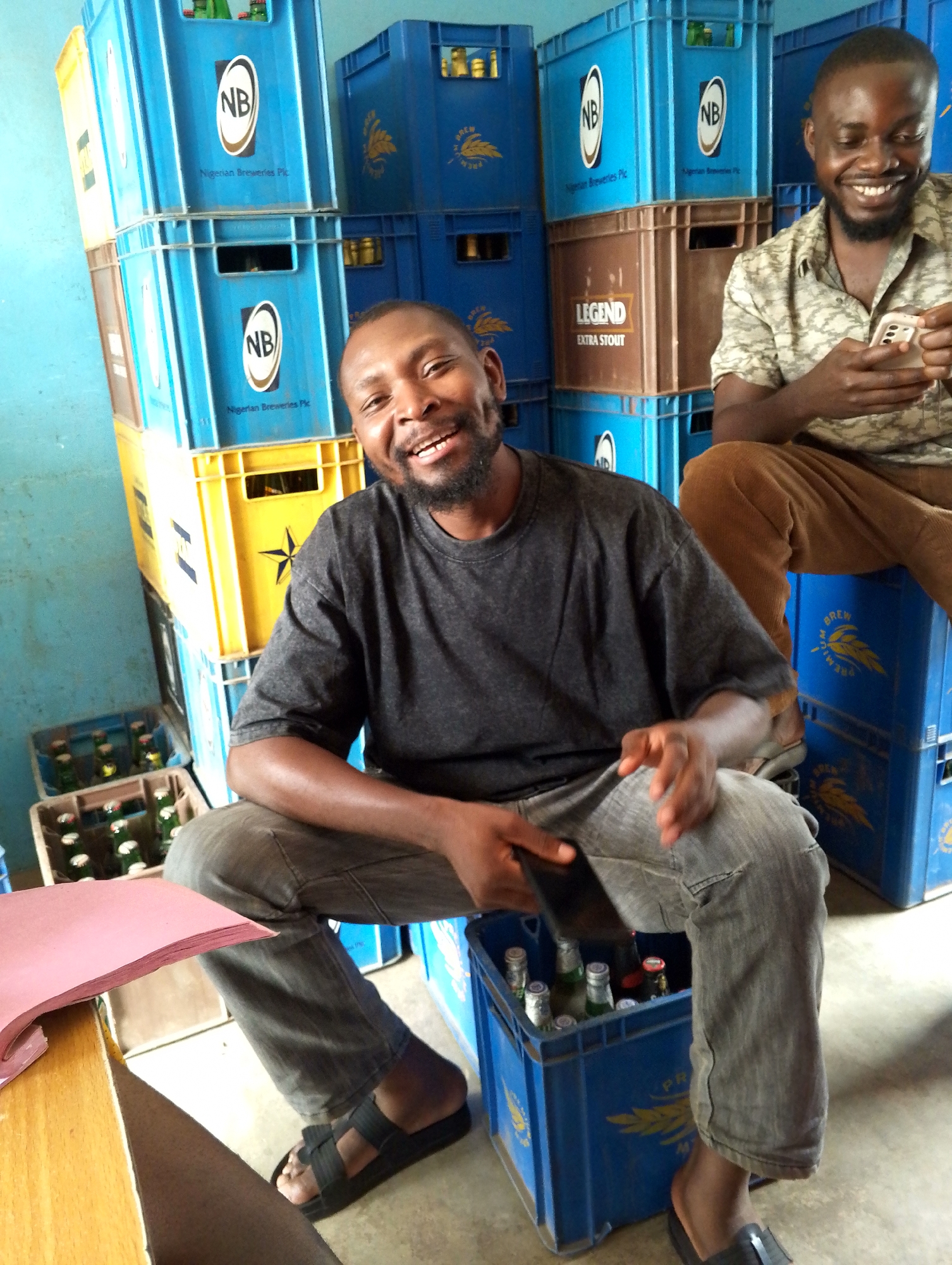 The image shows two men sitting amidst stacks of blue and yellow plastic crates, some labeled "NB Nigerian Breweries Plc," and one labeled "LEGEND EXTRA STOUT." The main subject, in the foreground, is wearing a gray t-shirt and gray jeans, along with black sandals. He is smiling and holding a black smartphone in his hands. He is sitting on a blue crate filled with beer bottles. The other man, in the background, is wearing a patterned shirt and brown pants. He is also smiling and looking at a white smartphone. The setting appears to be an informal outdoor or semi-outdoor area, possibly a small business or shop with a stack of beer crates. Part of a light-colored wooden desk or table is visible at the left edge of the frame.