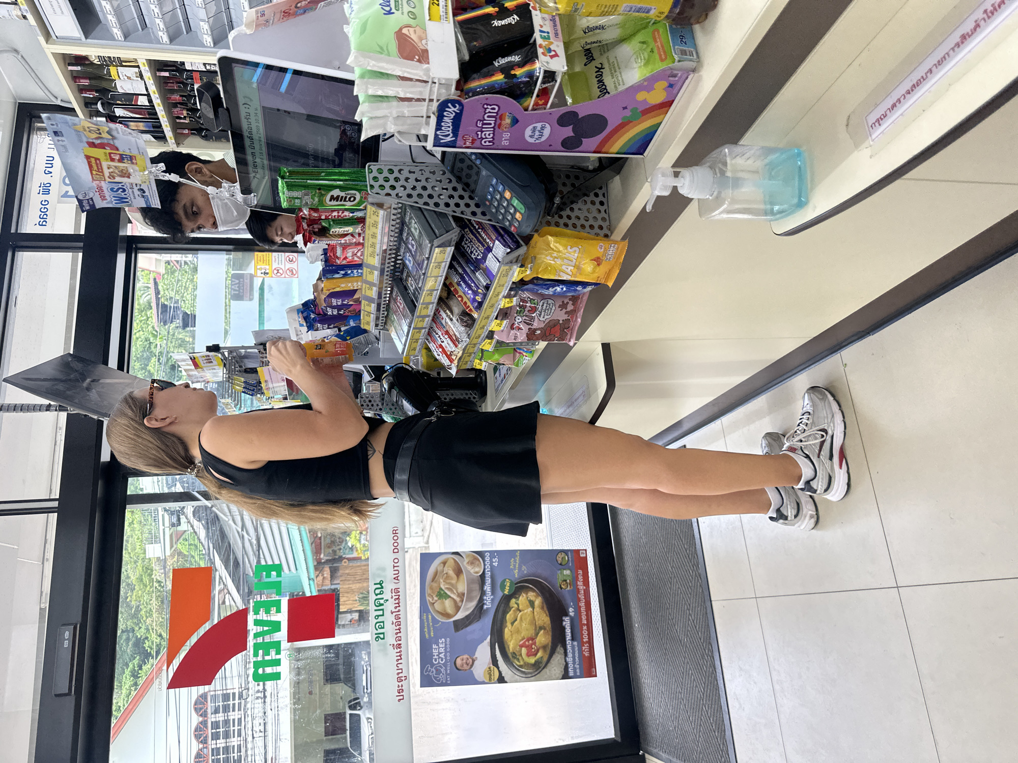 In Karon, Thailand, a young woman with long blonde hair tied in a ponytail, wearing a black tank top, a black belt, and a short black skirt, stands at a 7-Eleven checkout counter. She is looking to her right, holding an orange beverage in her left hand. In front of her, a male cashier, wearing a white face mask, is looking at a computer screen. The counter is stocked with various items, including candy, snacks, and toiletries. A hand sanitizer dispenser is on the counter to the right. The store has a tiled floor and a large window with the 7-Eleven logo visible on it, offering a view of a street and buildings outside.