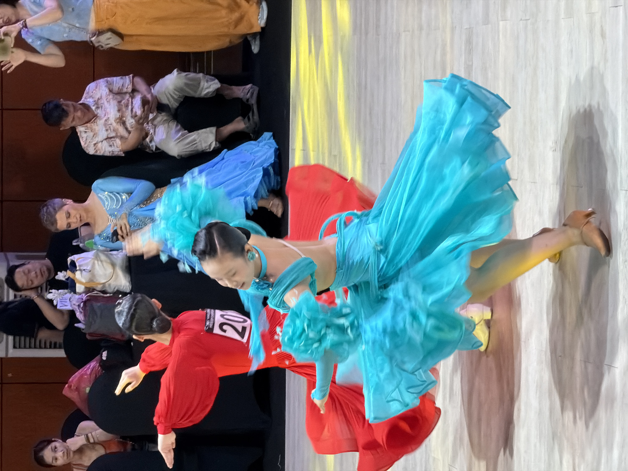In an indoor setting, likely a dance competition or performance venue in Hanoi, Vietnam, two young women are captured in motion, their brightly colored ballroom dance dresses creating a dynamic visual. The woman in the foreground, wearing a vibrant turquoise dress with voluminous ruffles and flowing sleeves, is the central focus. Her hair is styled in a sleek bun, and she wears a light-colored earring. Her body is angled away from the viewer, and her arms are extended, contributing to the sense of movement. The second dancer, to her left and slightly behind, is clad in a striking red dress with full sleeves, her back mostly to the camera. A participant number "20" is visible on her attire.
Surrounding the dancers, a blurred audience is seated, some individuals holding phones. The background features dark seating and a wooden wall. The floor is light-colored with a yellow light projection casting a colorful sheen. The lighting suggests an artificial environment, typical of event spaces. The overall atmosphere is one of active performance, with the dancers' dresses in mid-swirl, suggesting a moment of high energy within a choreographed routine.