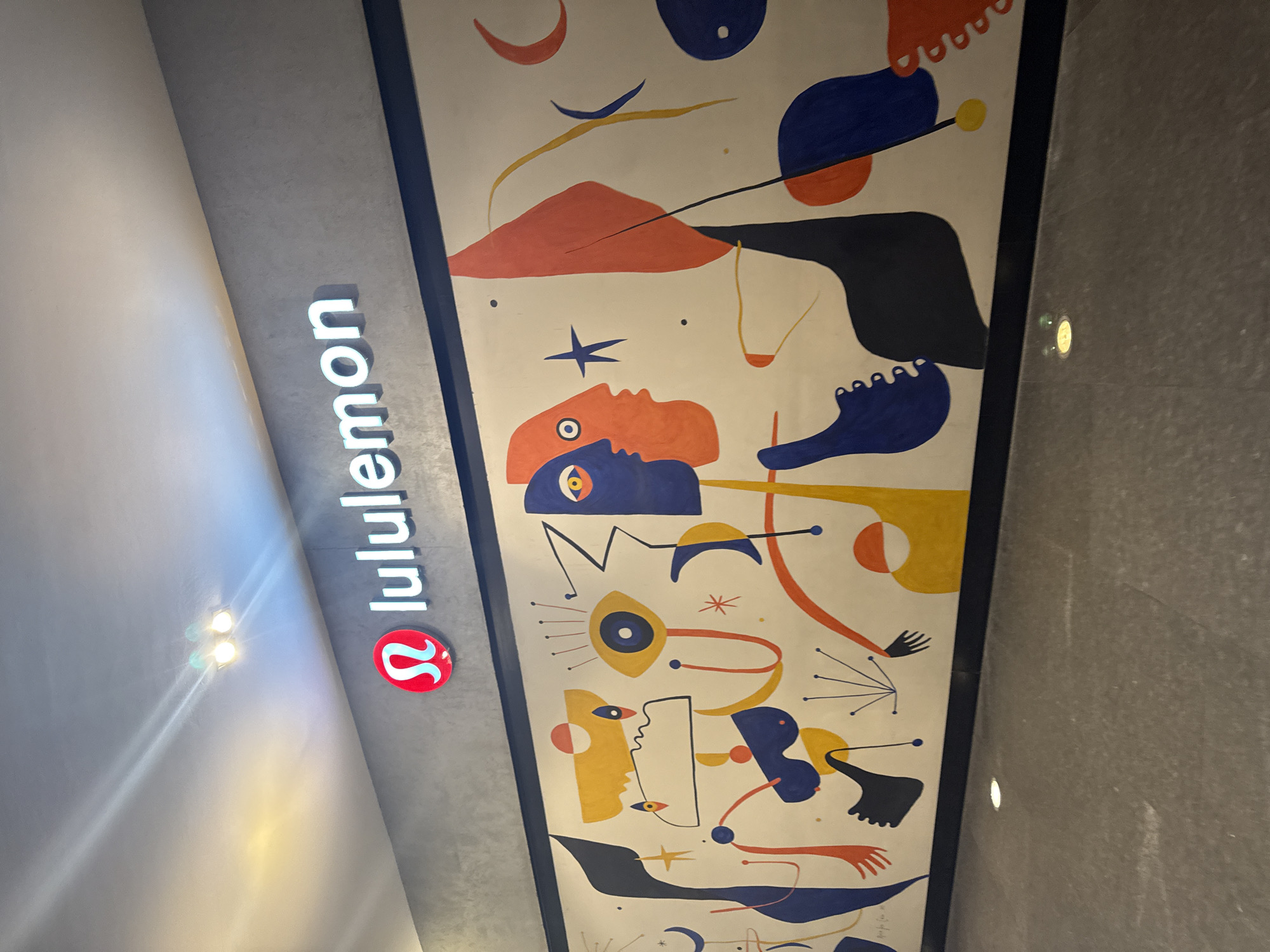 This media file captures an indoor view, likely within a modern commercial space such as a shopping mall or a retail store. The primary subjects are the Lululemon brand signage and a large, vibrant abstract mural.
The Lululemon signage is prominently displayed on a dark grey, textured wall. The word "lululemon" is spelled out in white, illuminated letters, positioned vertically along the wall. Below the text is the brand's distinctive red, circular logo featuring a stylized "A" shape in white. Bright overhead lights are visible, casting a glare on the upper left portion of the image and illuminating the sign.
Adjacent to the Lululemon signage is a tall, narrow abstract mural. The mural features a bold, colorful design in a style reminiscent of modern art, possibly inspired by artists like Joan Miró. The color palette includes vivid red-orange, various shades of blue, yellow, and black, set against a light, off-white background. The artwork consists of fragmented and stylized forms, including abstracted human-like figures, faces with prominent eyes, limbs, geometric shapes, stars, crescents, and dynamic lines and dots. The figures appear to be in various states of movement or interaction, creating a sense of energy and fluidity.
The overall setting suggests a contemporary and art-infused environment, likely a Lululemon store in Puebla, Mexico, given the provided context. The activity captured is merely a visual observation of the store's branding and artistic decor. The time of day cannot be determined, but the indoor lighting indicates a well-lit space.