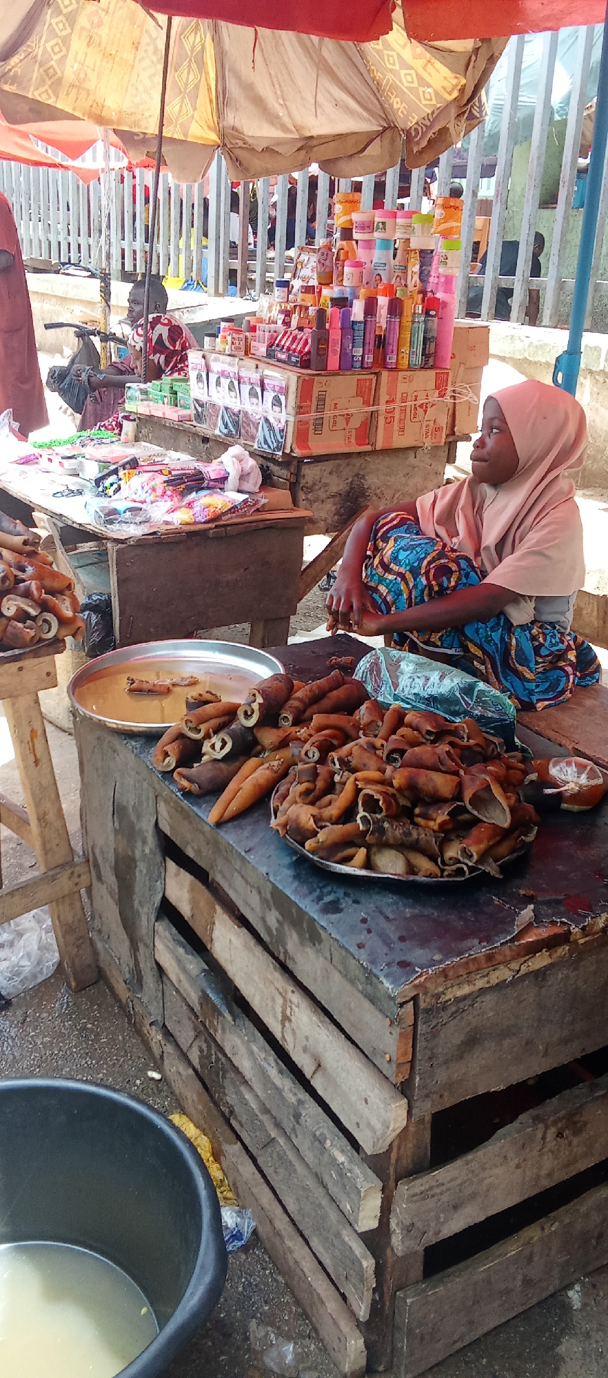 A street vendor sits behind a table laden with cooked cow skin, known as "ponmo," for sale at a market in Akwanga, Nigeria. She is a young girl wearing a light tan hijab and a blue and brown patterned wrapper. Her posture is relaxed as she looks off to the side. The ponmo is displayed on metal platters. Other merchandise, including cosmetics, hair products, and hair accessories, are arranged on tables behind her. A large black bucket filled with a light-colored liquid sits at the lower left of the frame. The tables are wooden and worn, suggesting a well-used market stall. The scene is lit by bright sunlight filtering through a large, worn umbrella that covers the stall. A metal fence is in the background. Another person sits at a nearby stall.