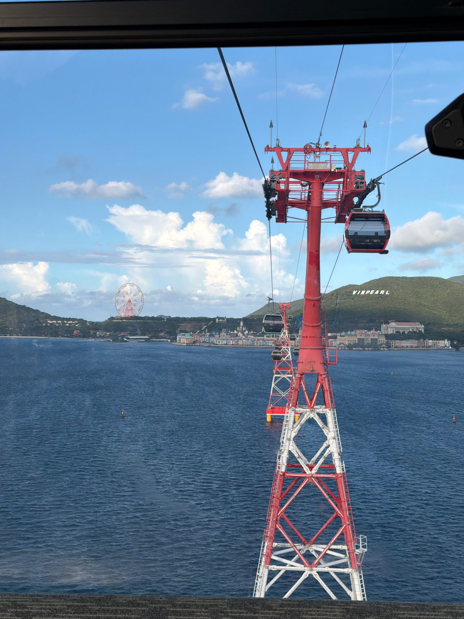 The image captures a view from a cable car in Nha Trang, Vietnam, overlooking the ocean and a coastal city. In the foreground, a red and white cable car tower rises from the water, supporting multiple cable cars suspended on wires. One red and black cable car is prominently positioned near the top of the tower. In the middle ground, the blue ocean stretches out, with a few buoys visible. Further in the distance, a Ferris wheel stands out against the skyline of a city built along the coast. The city features numerous buildings with red roofs and pastel-colored facades, suggesting a resort or entertainment complex. Behind the city, a lush green hill rises, with the word "VINPEARL" emblazoned in white letters on its slope. The sky above is a clear blue, dotted with fluffy white clouds, indicating fair weather. The overall scene depicts a popular tourist destination with a scenic coastal landscape and amusement attractions.